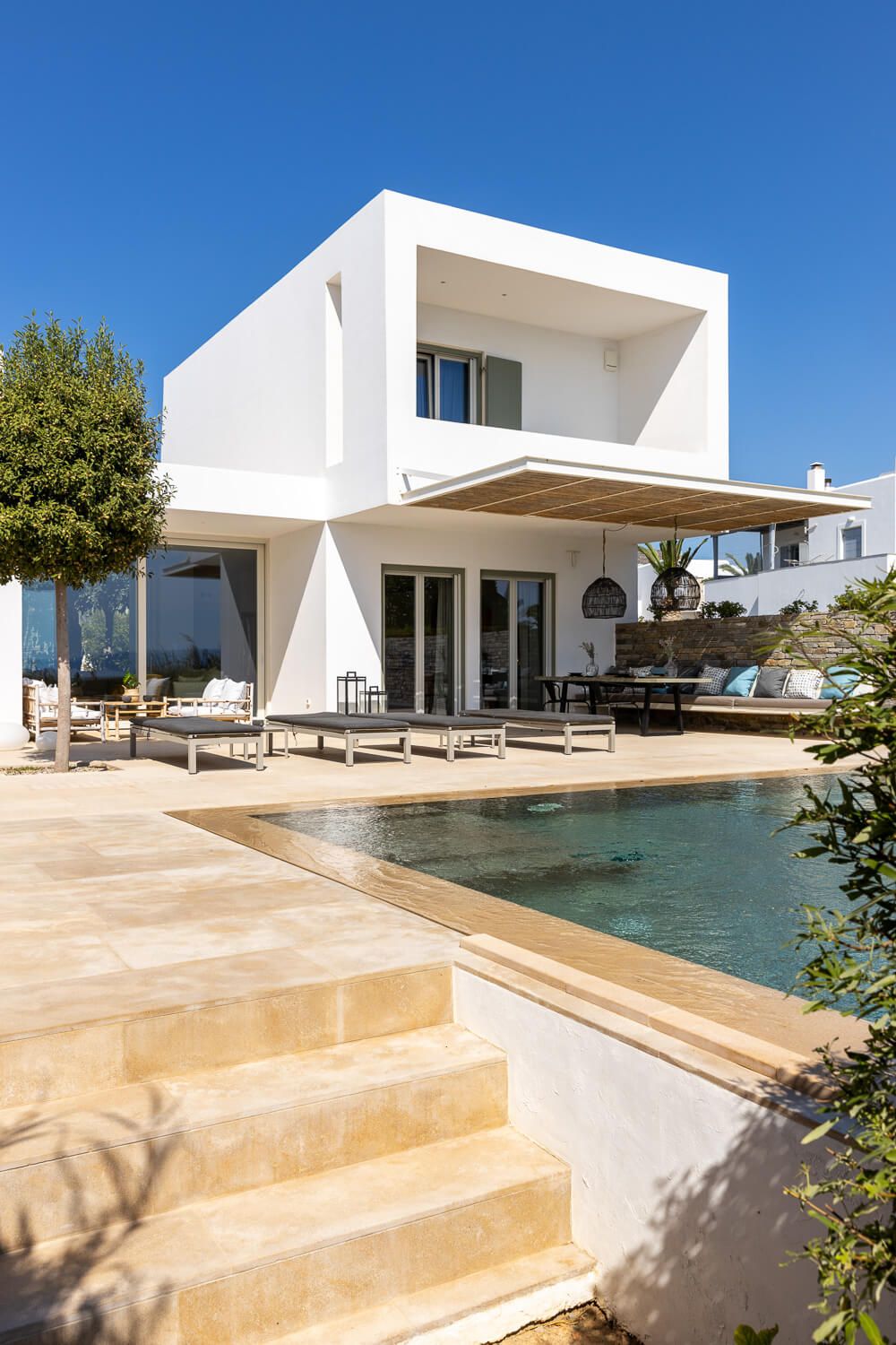 Modern luxury villa with pool, outdoor seating area, and stone steps under a blue sky in Cyclades.