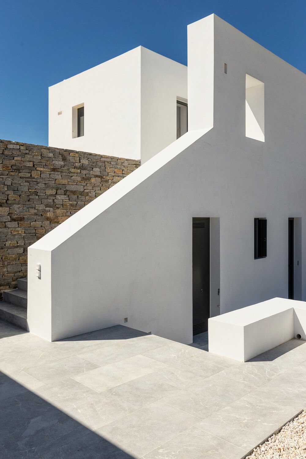 White minimalist Mediterranean architecture with staircase, stone wall accent under clear blue sky.