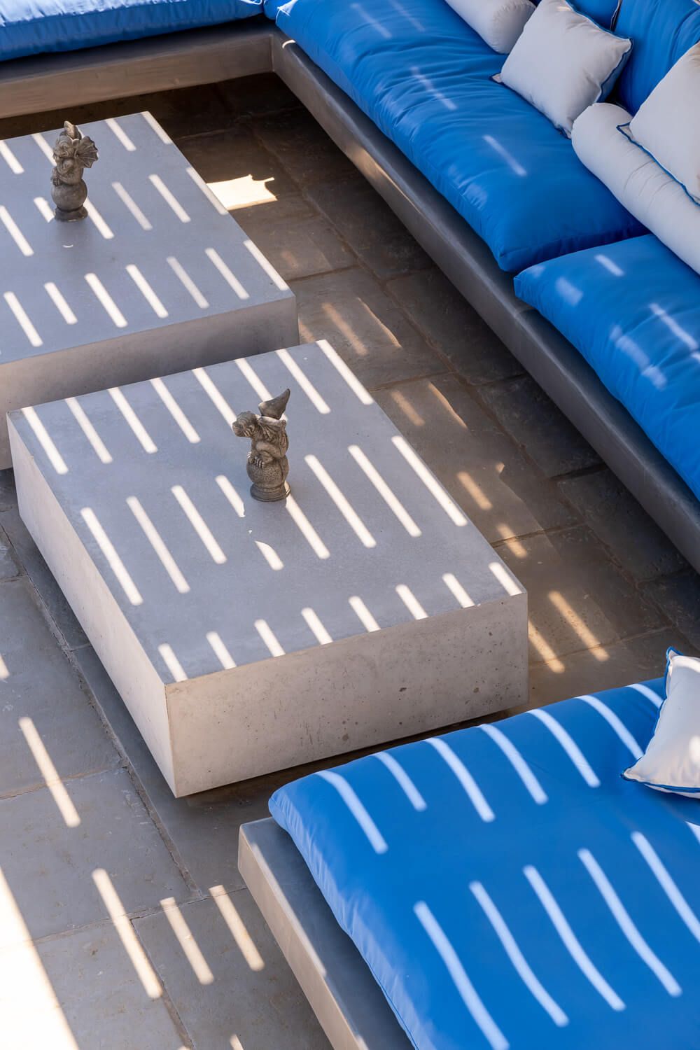 Modern outdoor seating area with bright blue cushions and concrete tables casting dramatic striped shadows.