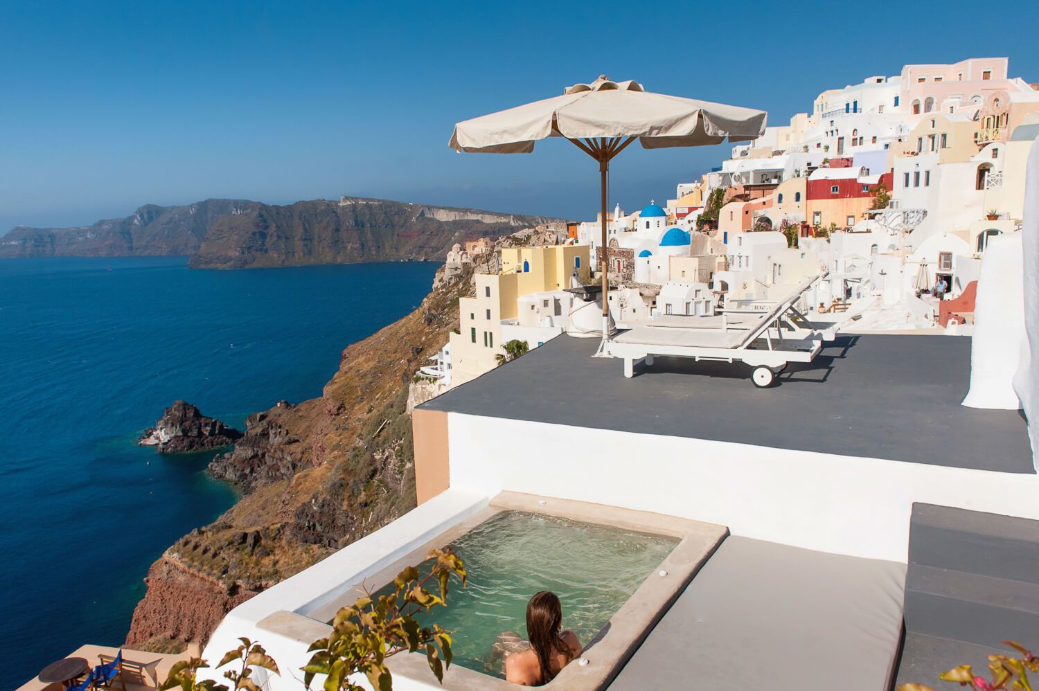 Woman relaxing in rooftop hot tub overlooking Santorini caldera and blue Aegean Sea views.