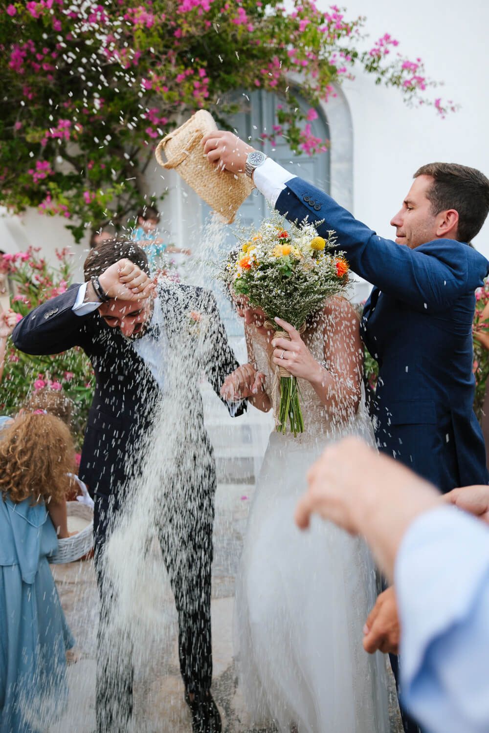 Wedding guests shower bride and groom with rice outside white church adorned with pink flowers.