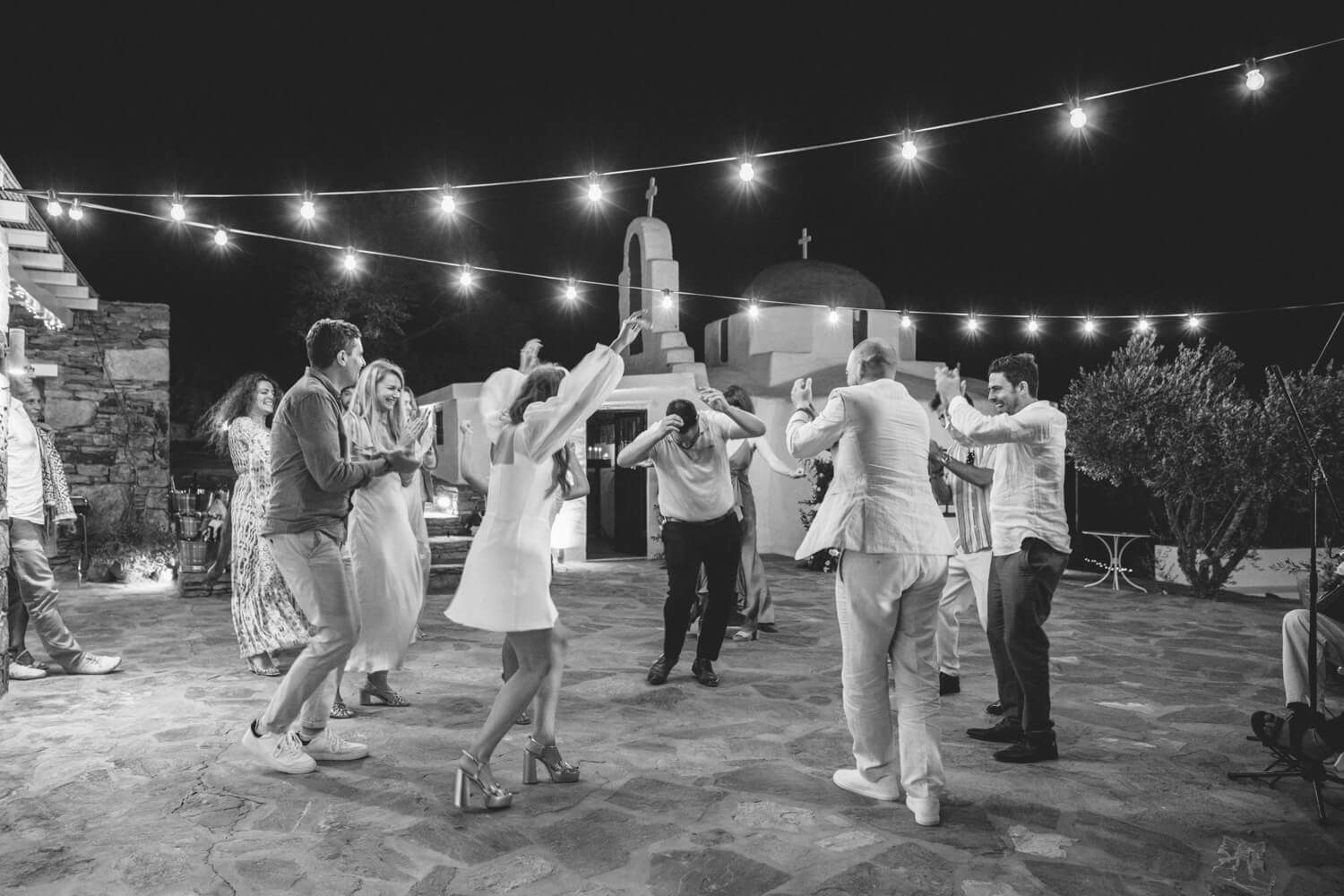 Black and white photo of wedding guests dancing in circle under string lights at night.