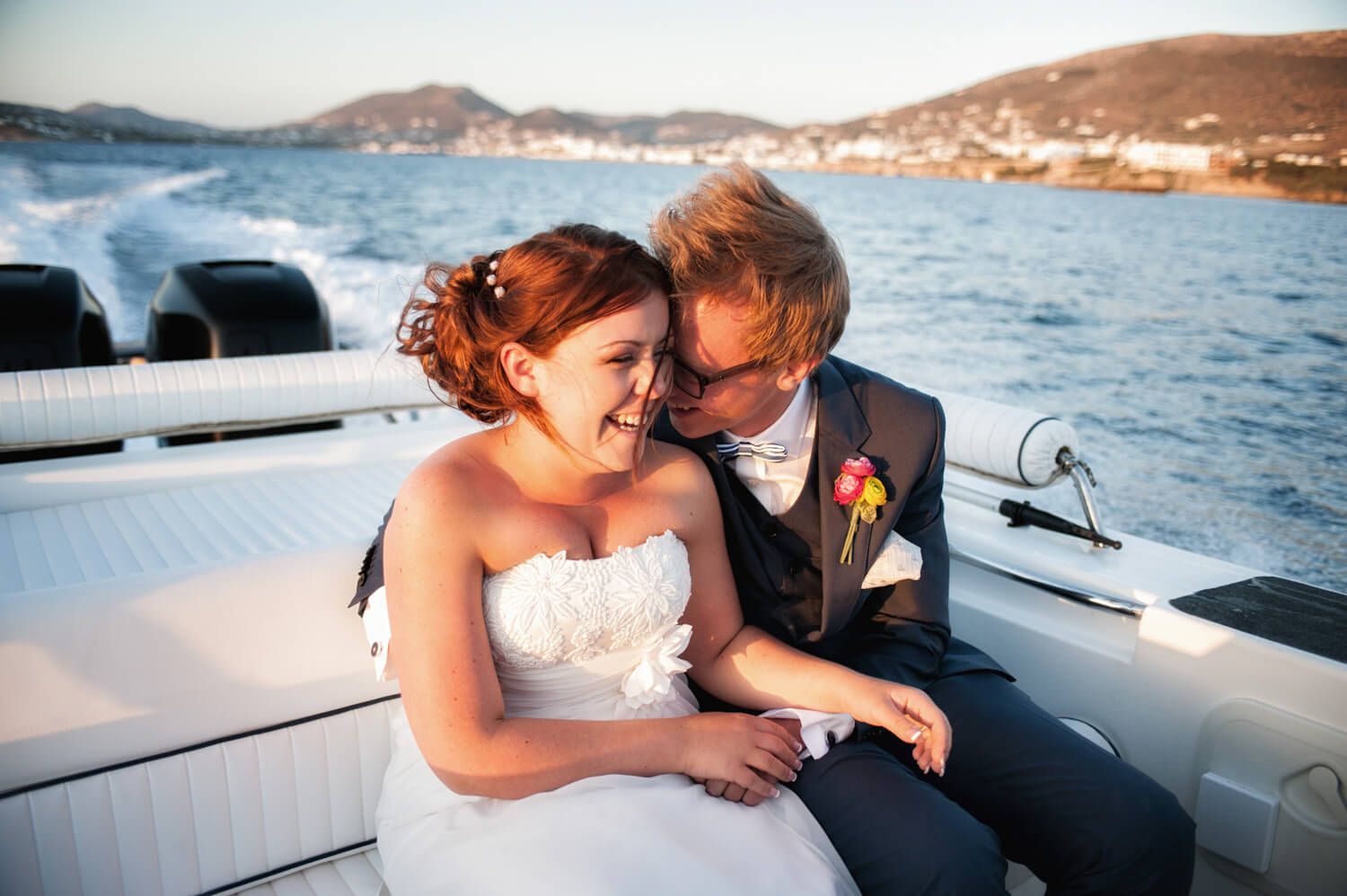 Newlywed couple sharing a romantic moment on a speedboat with ocean and hills in the background.