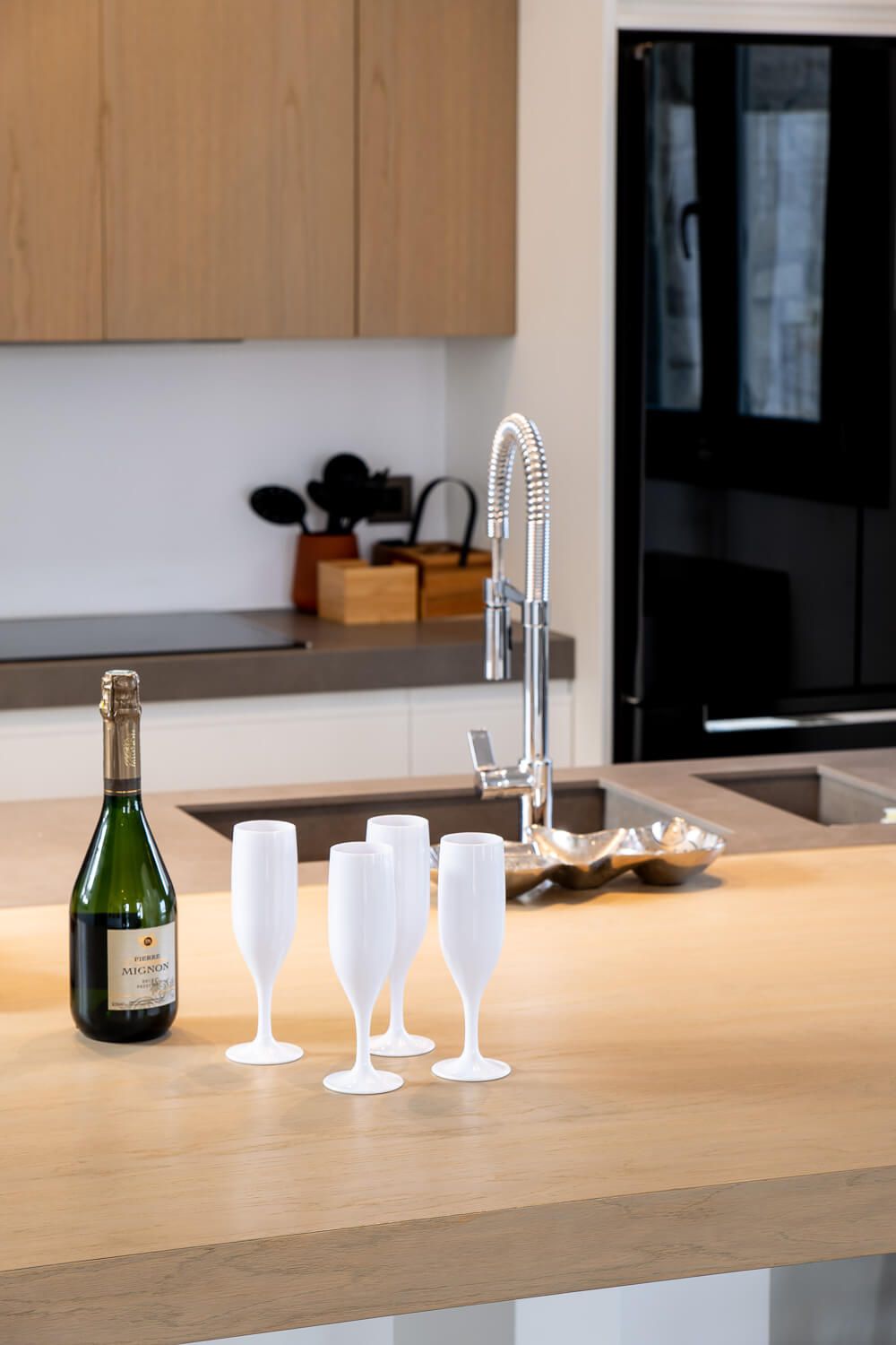 Modern kitchen counter with champagne bottle, frosted white glasses and sleek faucet against wooden cabinets and black appliances.