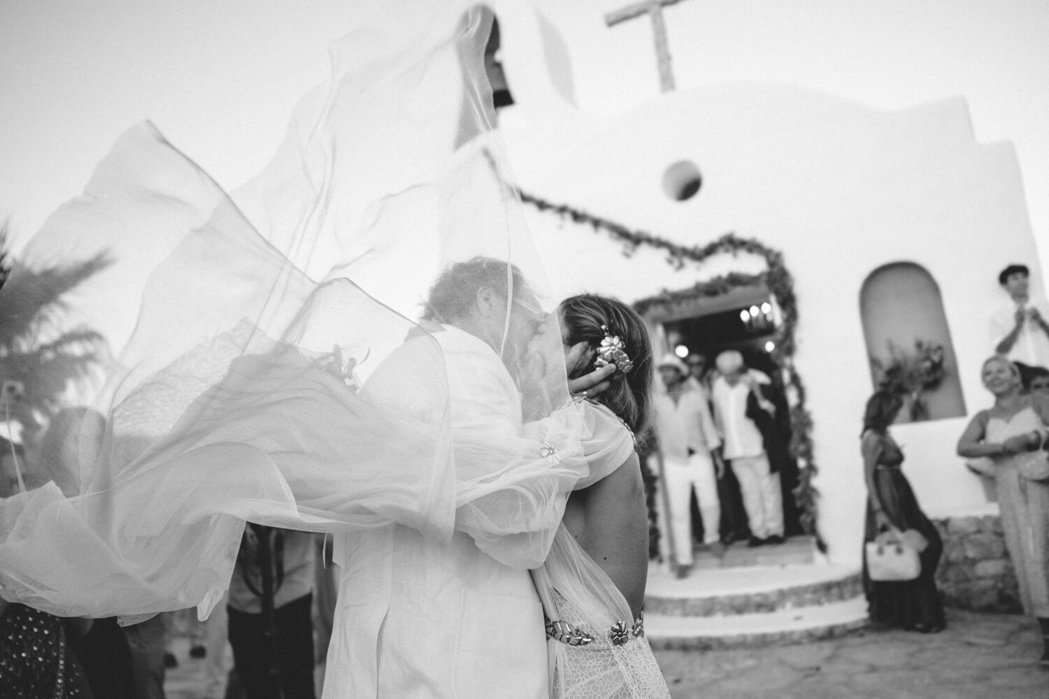 Bride's veil blowing in the wind outside a whitewashed Greek chapel during a wedding ceremony.