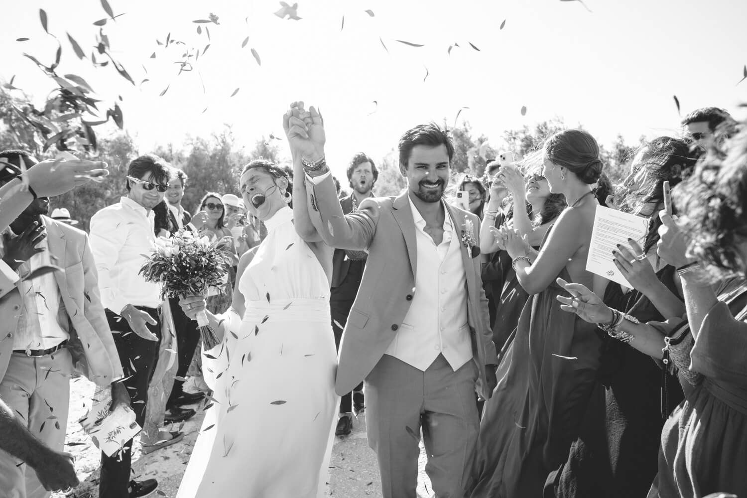 Black and white photo of newlyweds walking through confetti shower with cheering wedding guests.