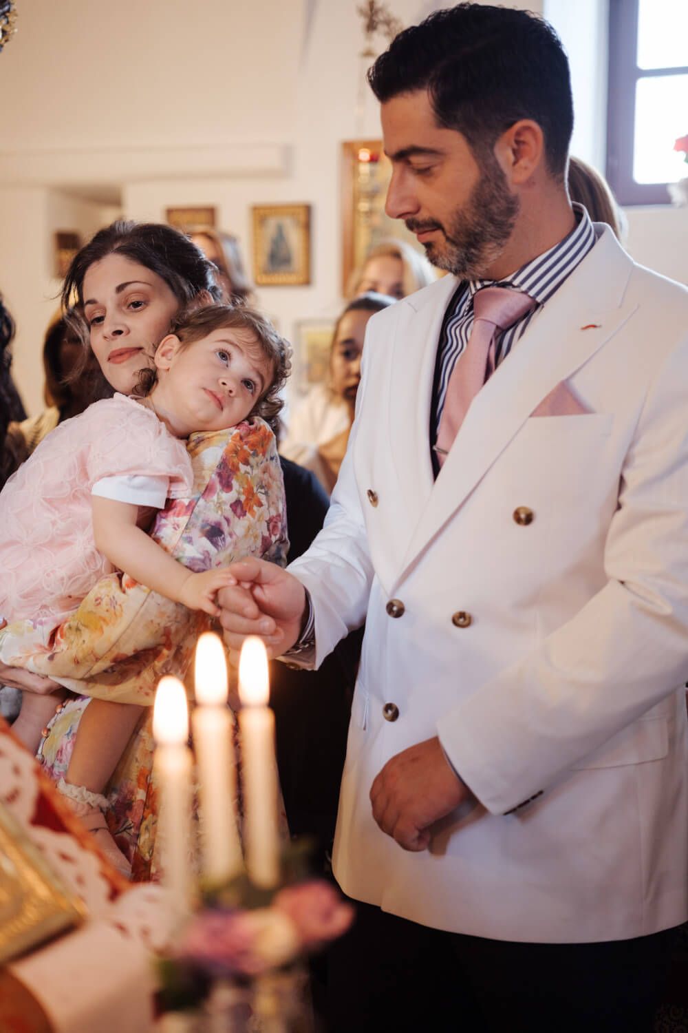 Man in white suit holds child's hand near candles during Orthodox baptism ceremony.