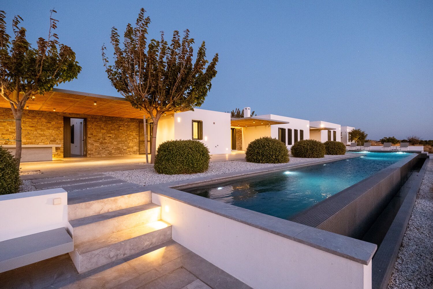 Luxury modern villa with illuminated lap pool, manicured hedges, and stone facade at dusk.
