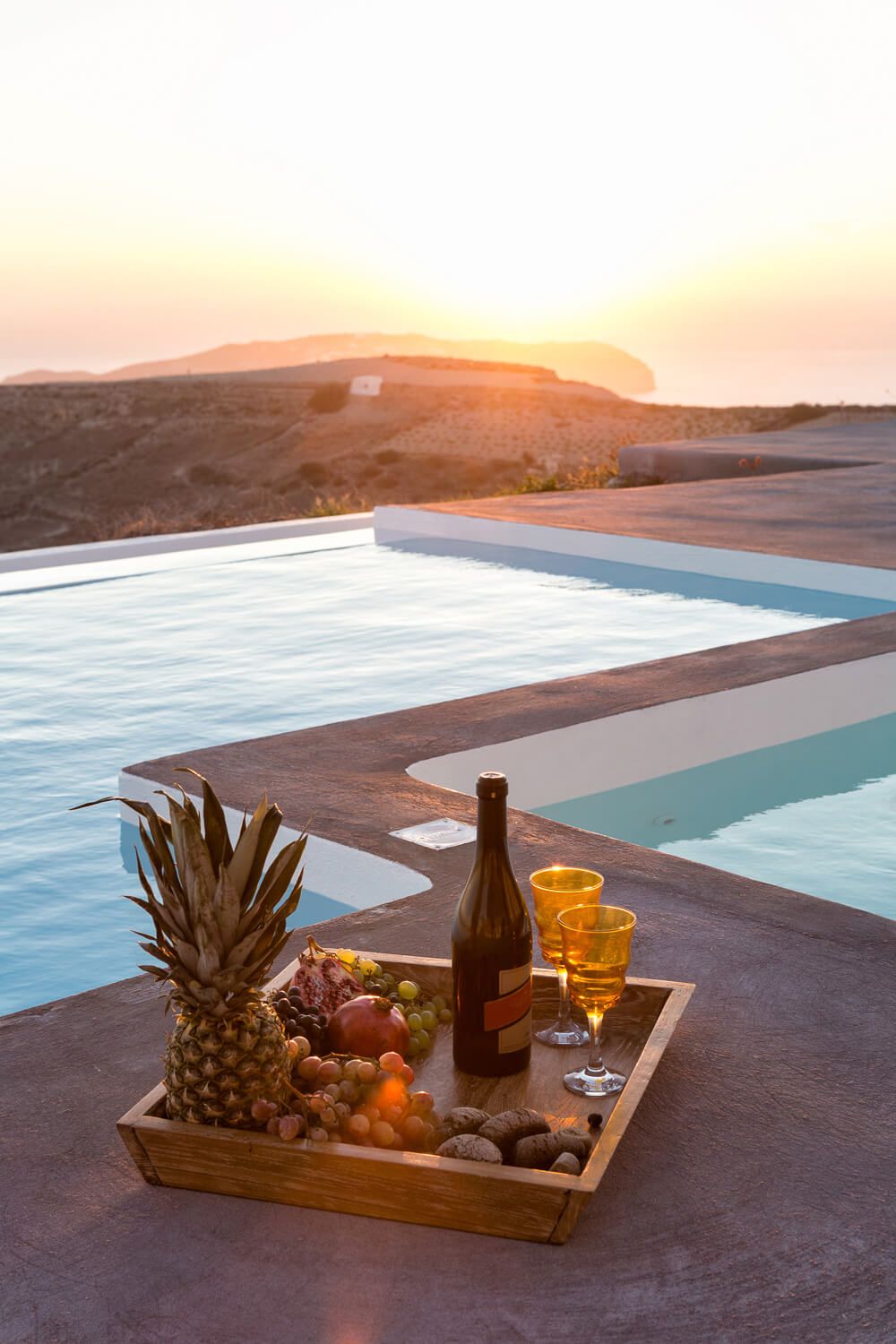 Tray with wine, fruit, and nuts beside an infinity pool at sunset overlooking the sea of Santorini.
