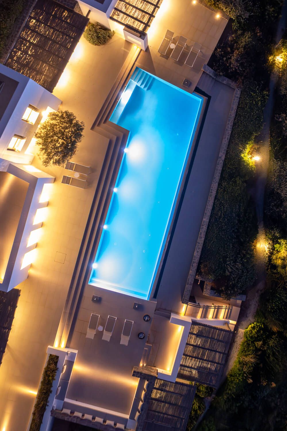 Aerial view of an illuminated swimming pool at night with warm lighting around the edges and blue water glowing.