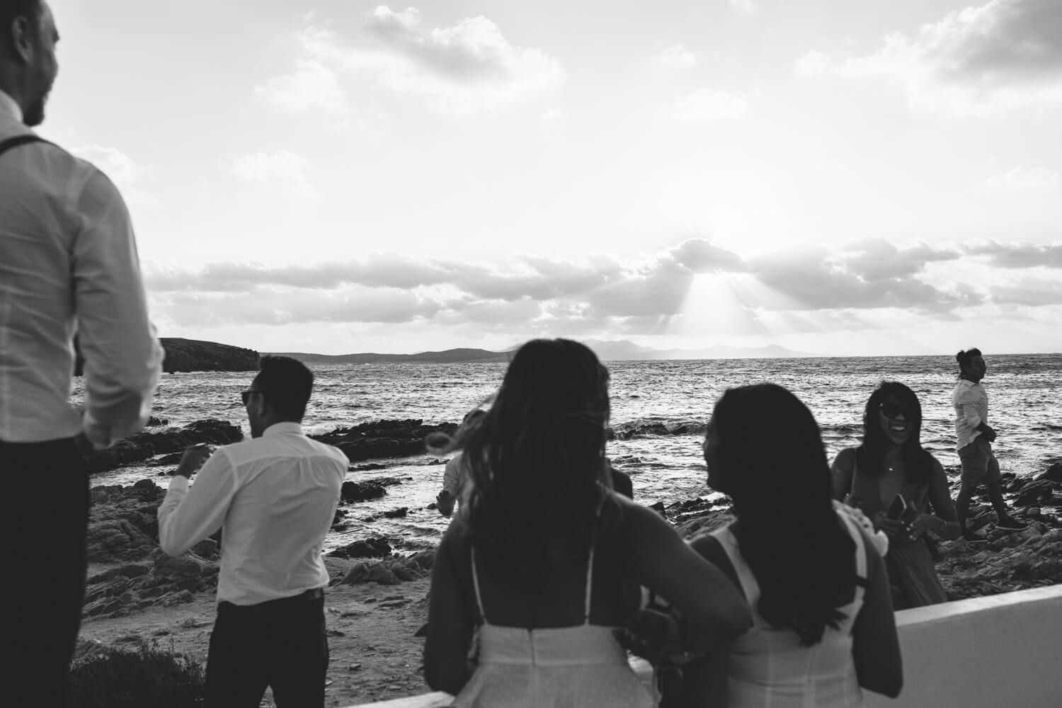 Wedding guests watching sunset over ocean from coastal venue in black and white photography.