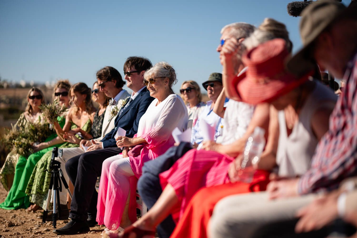 A crowd of people sitting in chairs outdoors at a wedding ceremony venue during daytime.