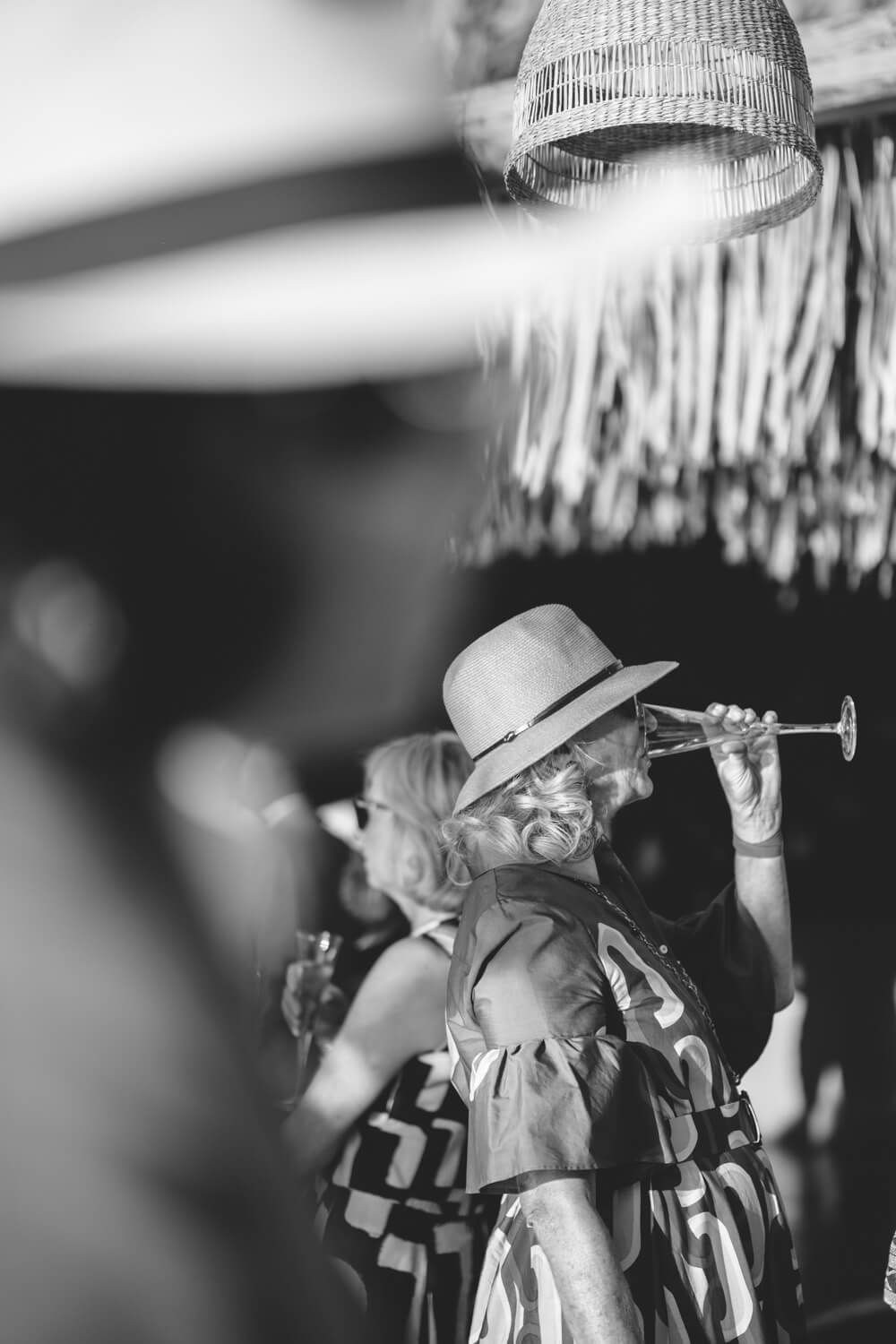 Black and white close-up of wedding guest in hat drinking from small trumpet-shaped glass.