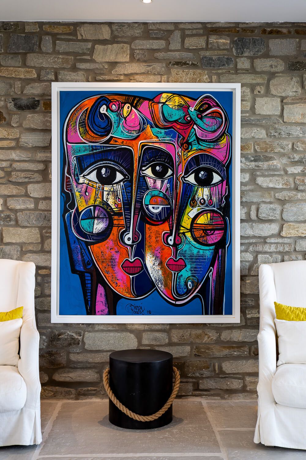 Modern abstract art prints featuring vibrant faces and geometric shapes hang on a stone wall above white chairs.
