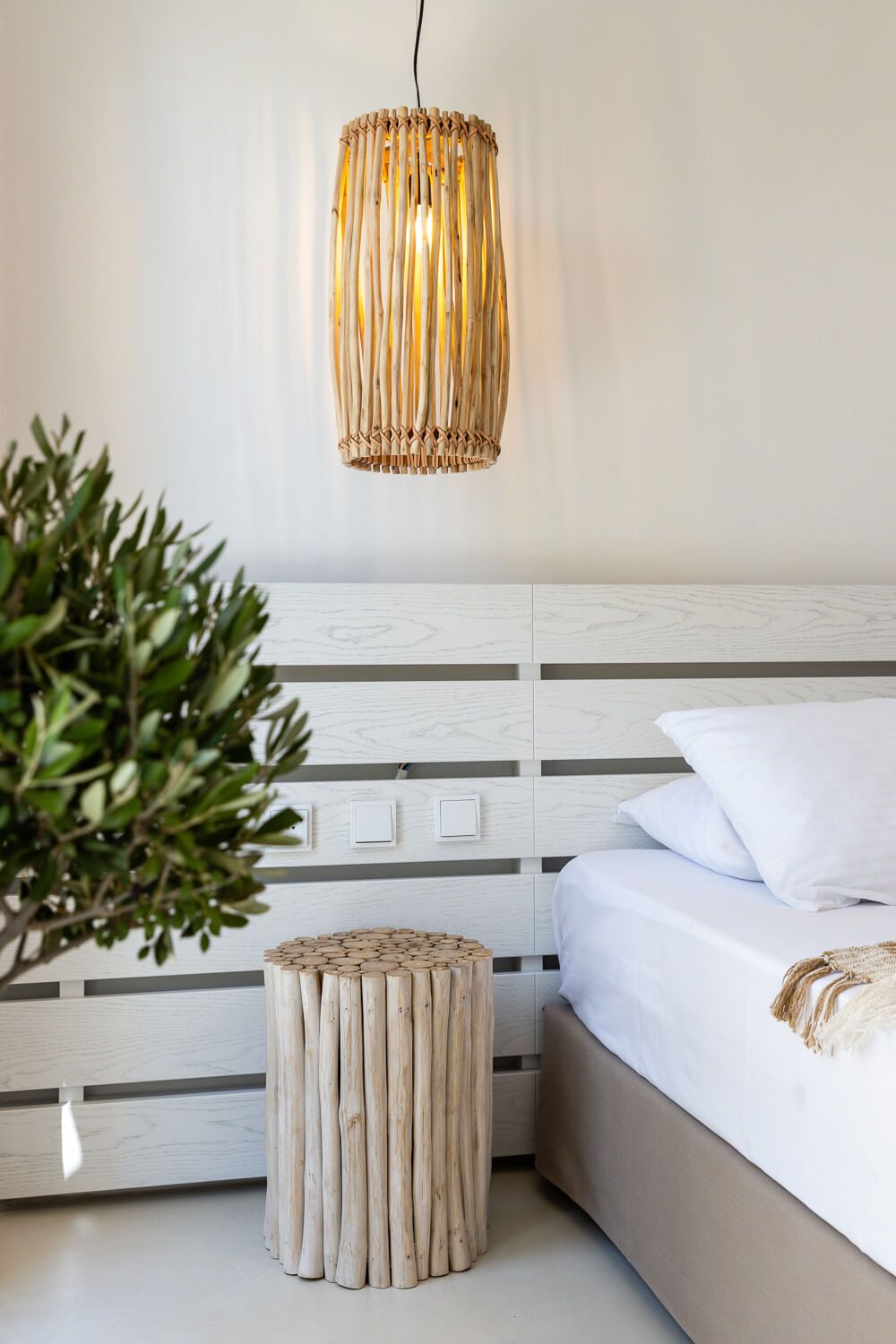 Minimalist bedroom interior with white bedding, natural driftwood side table, and rattan pendant light fixture.
