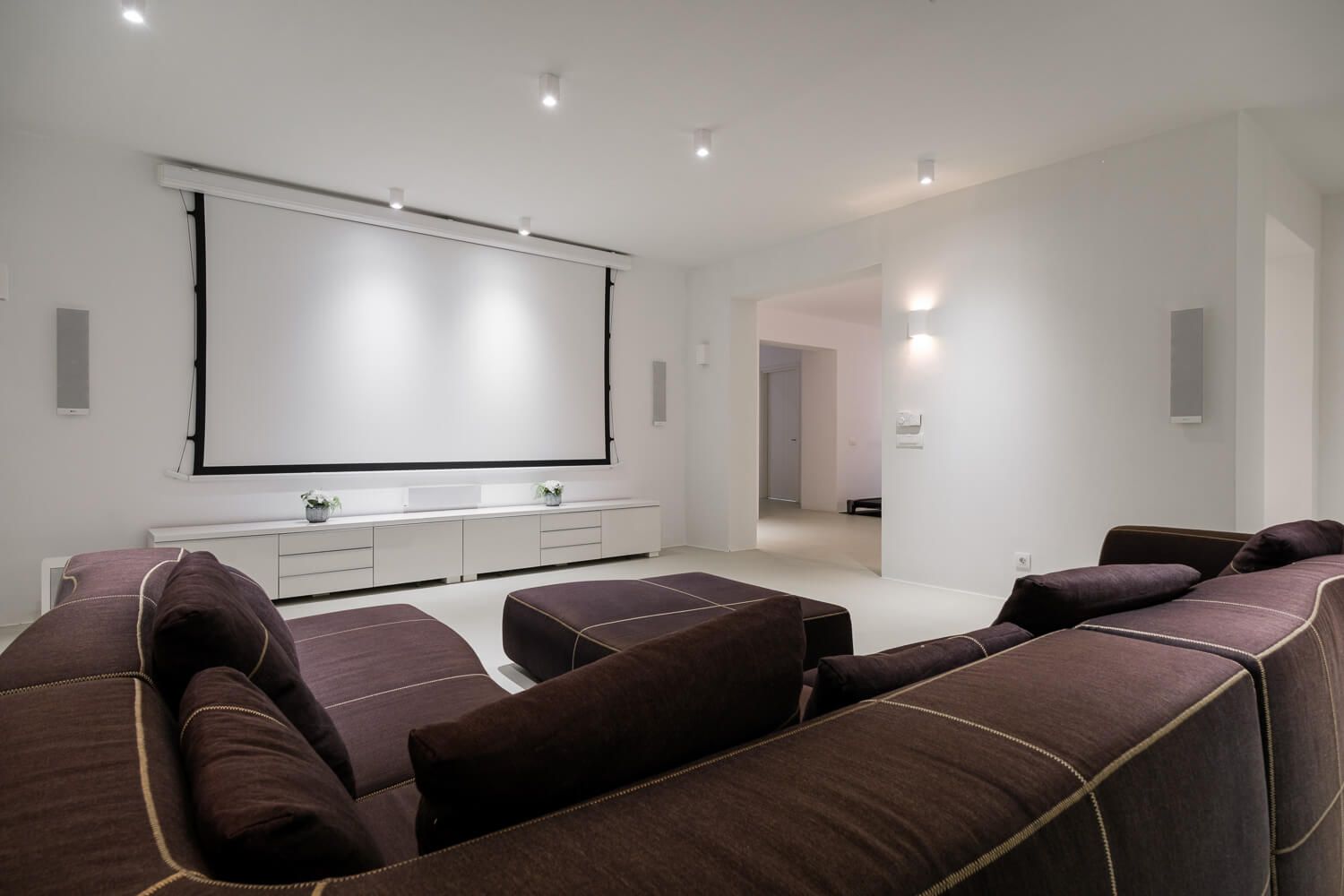 Modern home theater room with brown sectional sofa and large projector screen mounted on white wall.