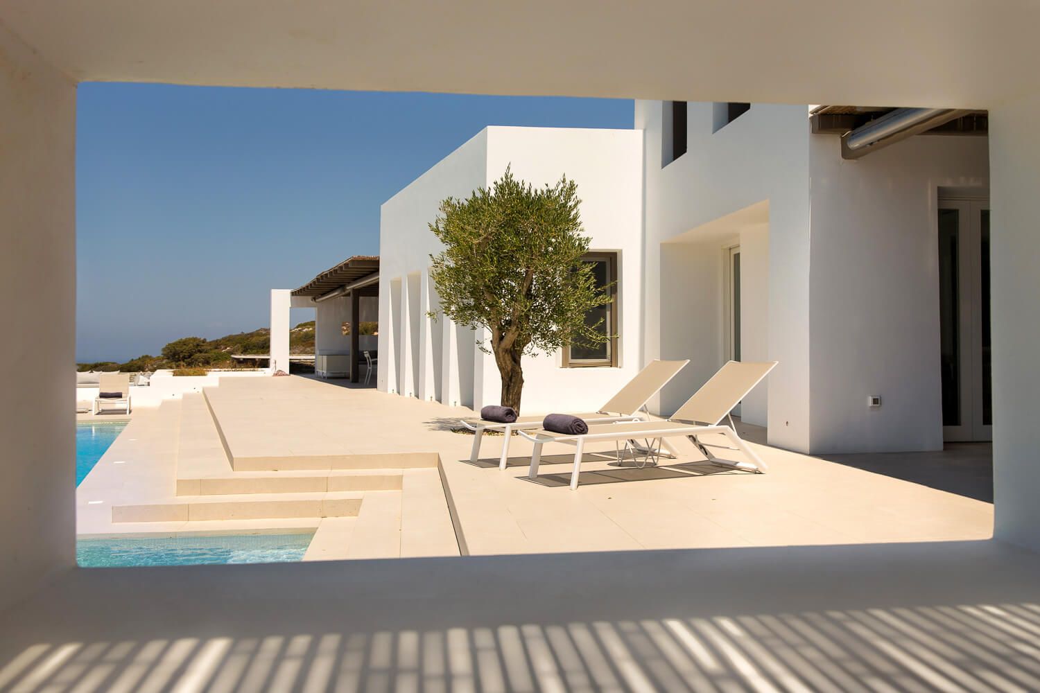 Modern white Mediterranean villa with sun loungers and olive trees on a sunny terrace with pool.