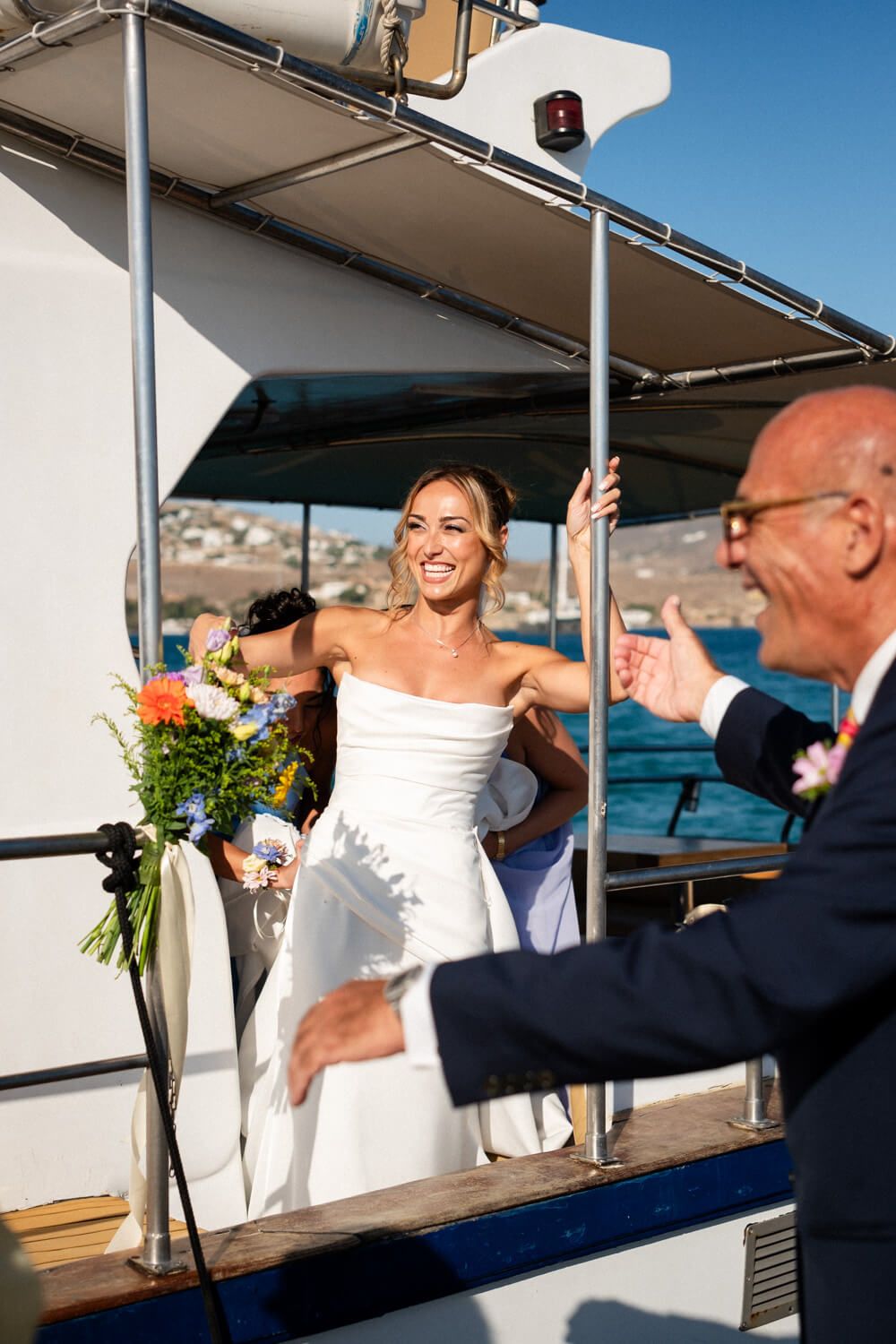 Radiant bride in strapless white gown smiling and waving from a yacht on her wedding day.