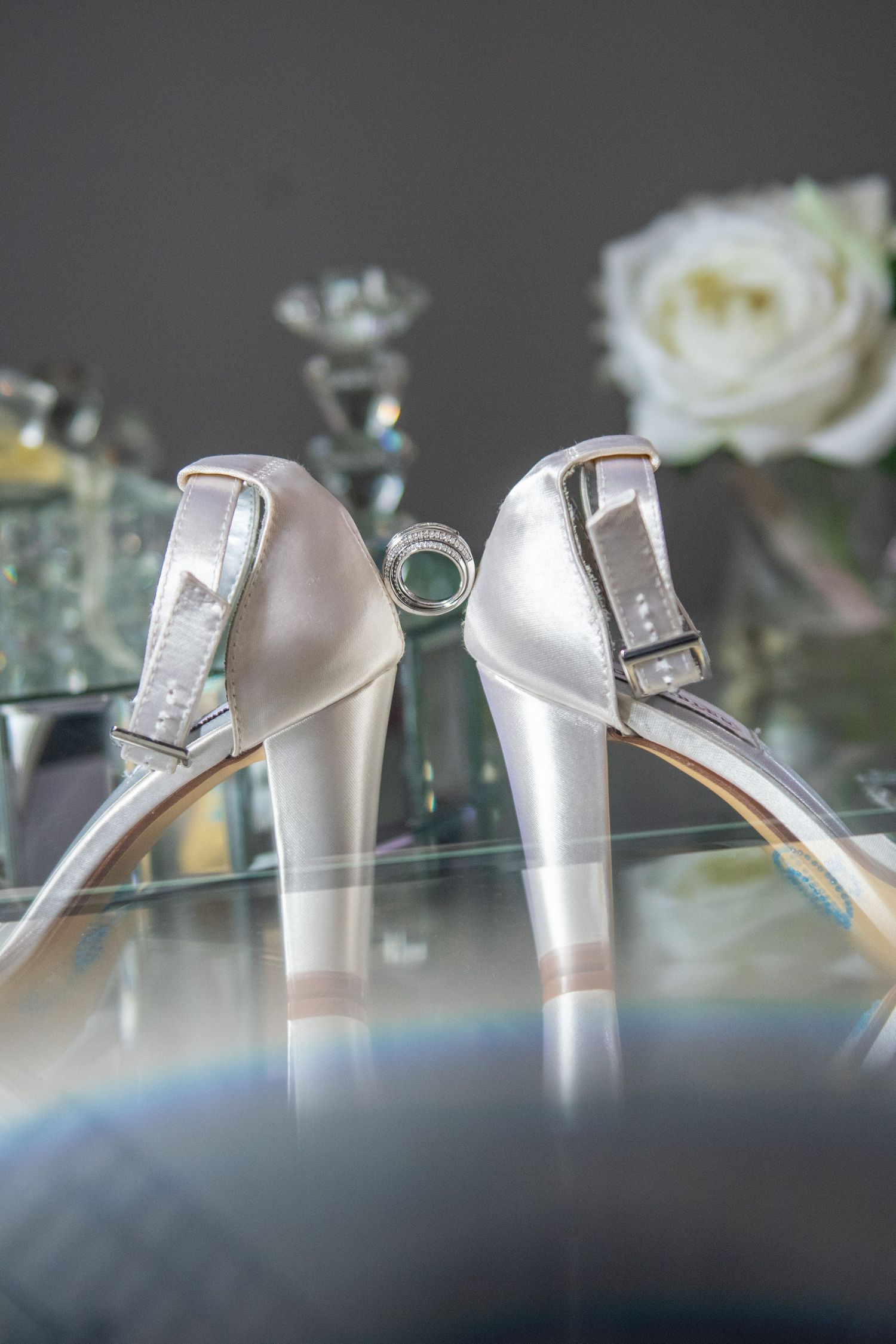 White satin wedding heels with ring photographed against reflective surface.