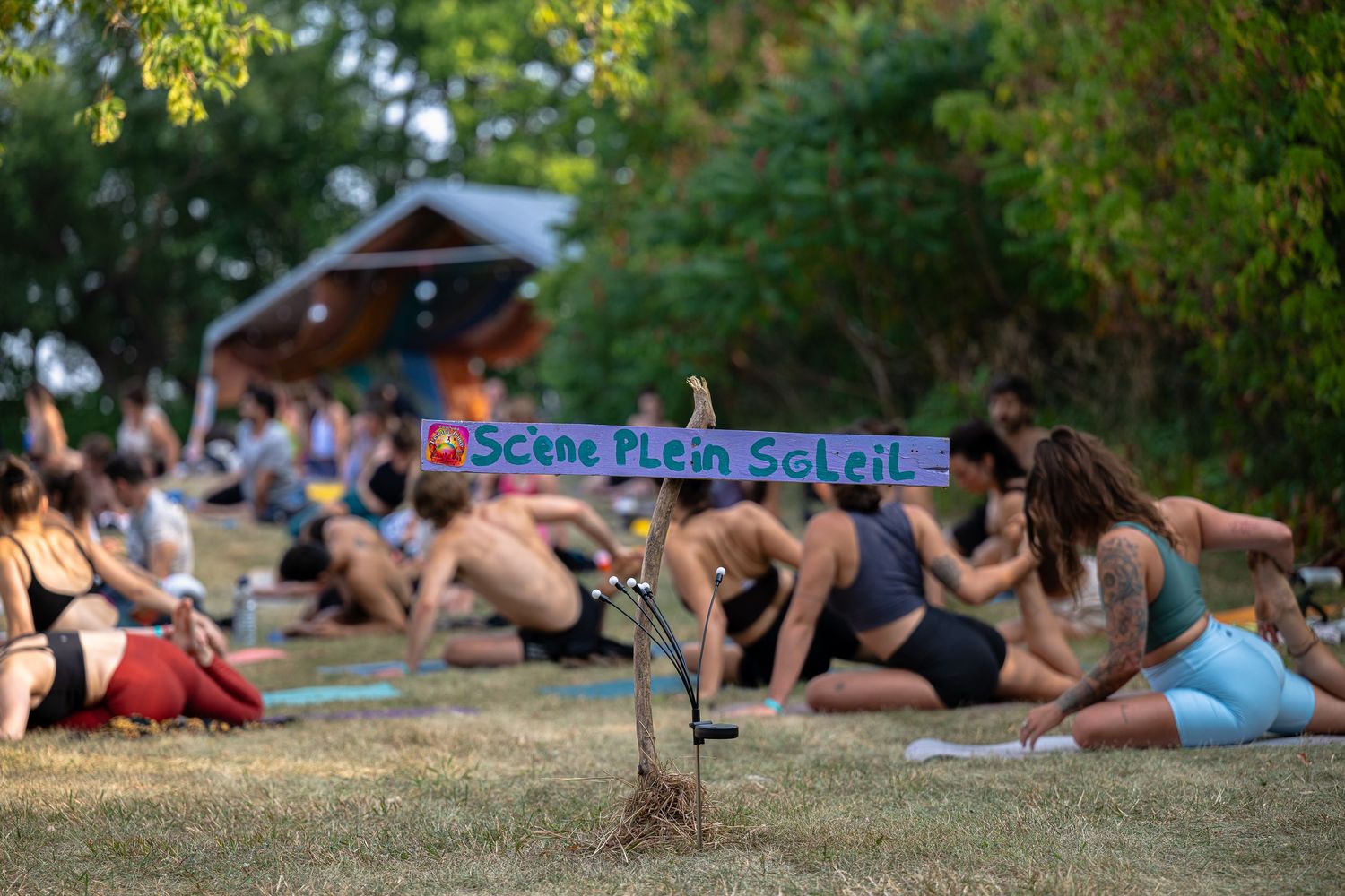 La structure en bois portant l’inscription 'Scène Plein Soleil' se dresse à l’Anatha Fest à Montréal, où un groupe pratique le yoga.