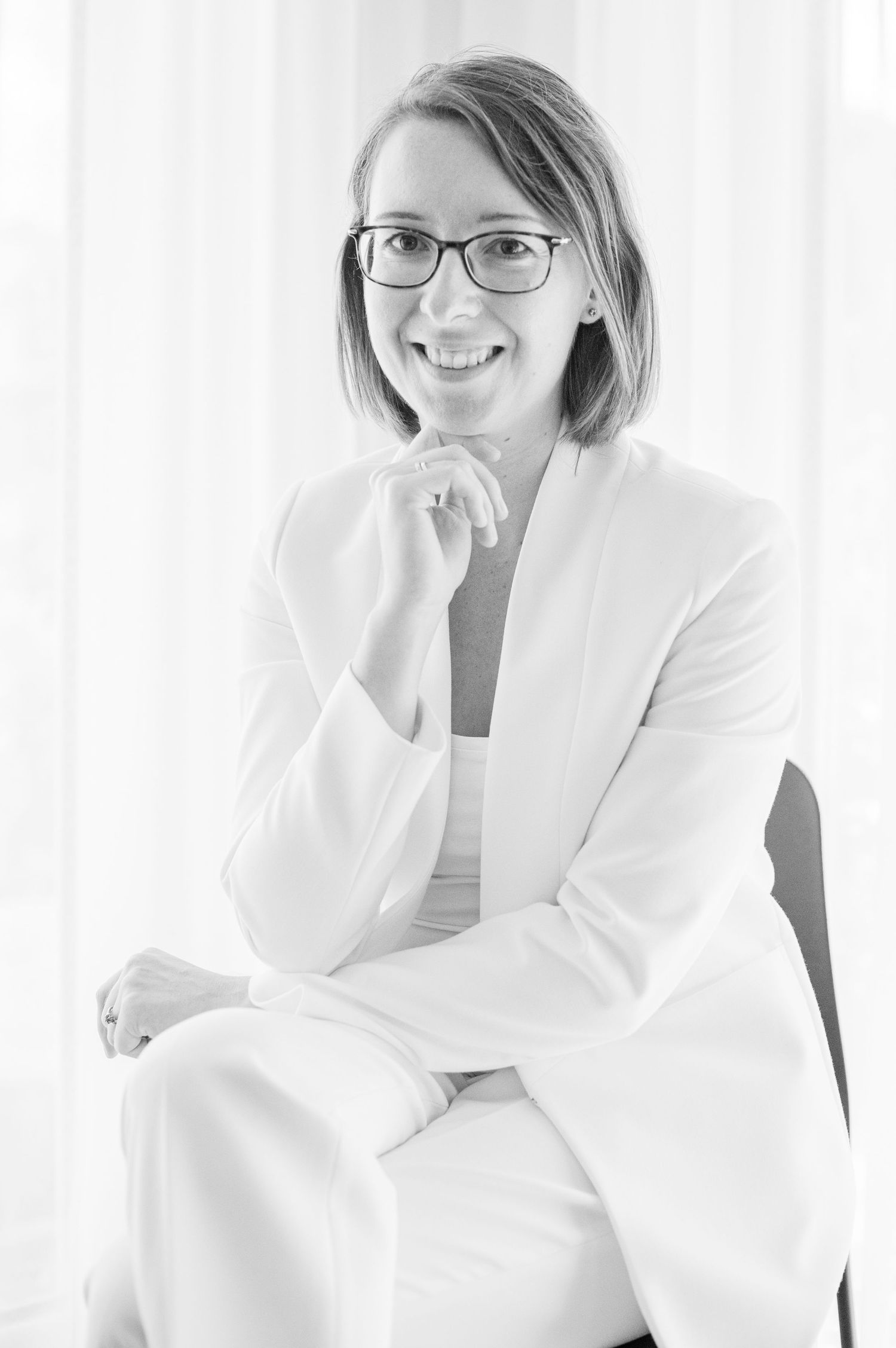 Ein professionelles Porträtfoto in Schwarz-Weiß der Fotografin Verena Frank, mit Brille und weißem Hosenanzug, sitzend mit der Hand am Kinn