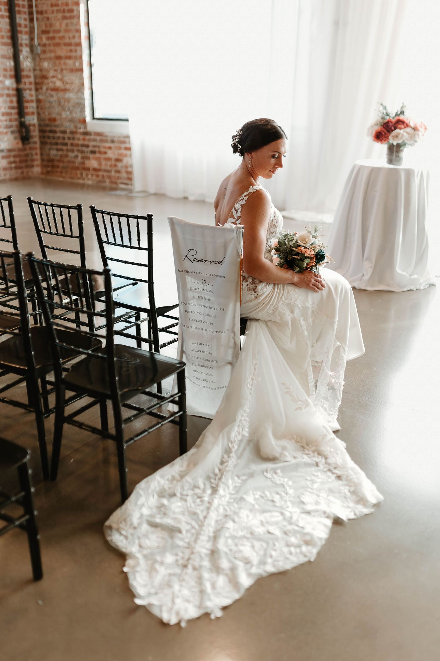 Bride in lace wedding gown with train holds bouquet beside reserved sign at elegant indoor ceremony venue.