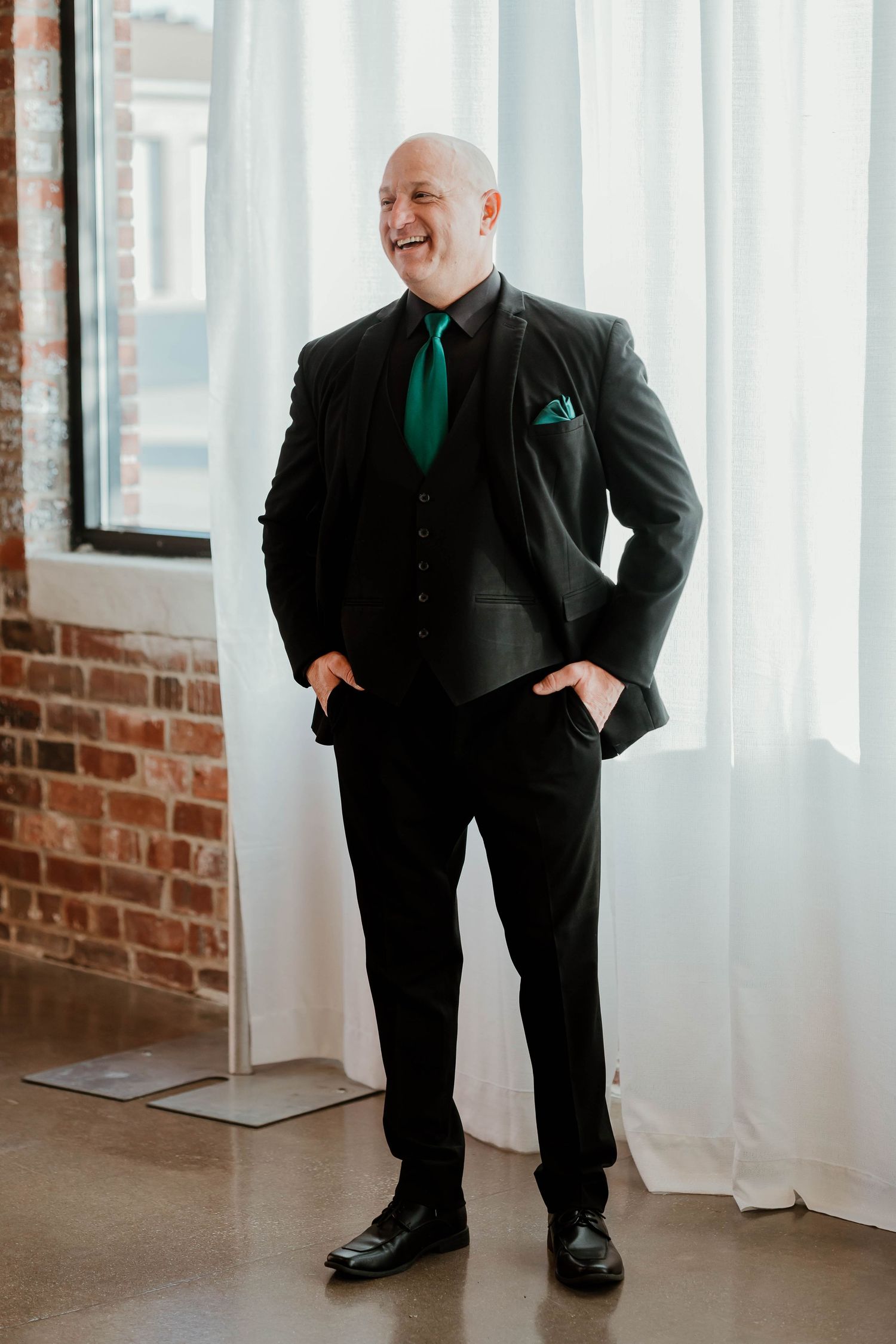 Bald man in black suit with green tie and pocket square, smiling confidently in a bright room with exposed brick wall.