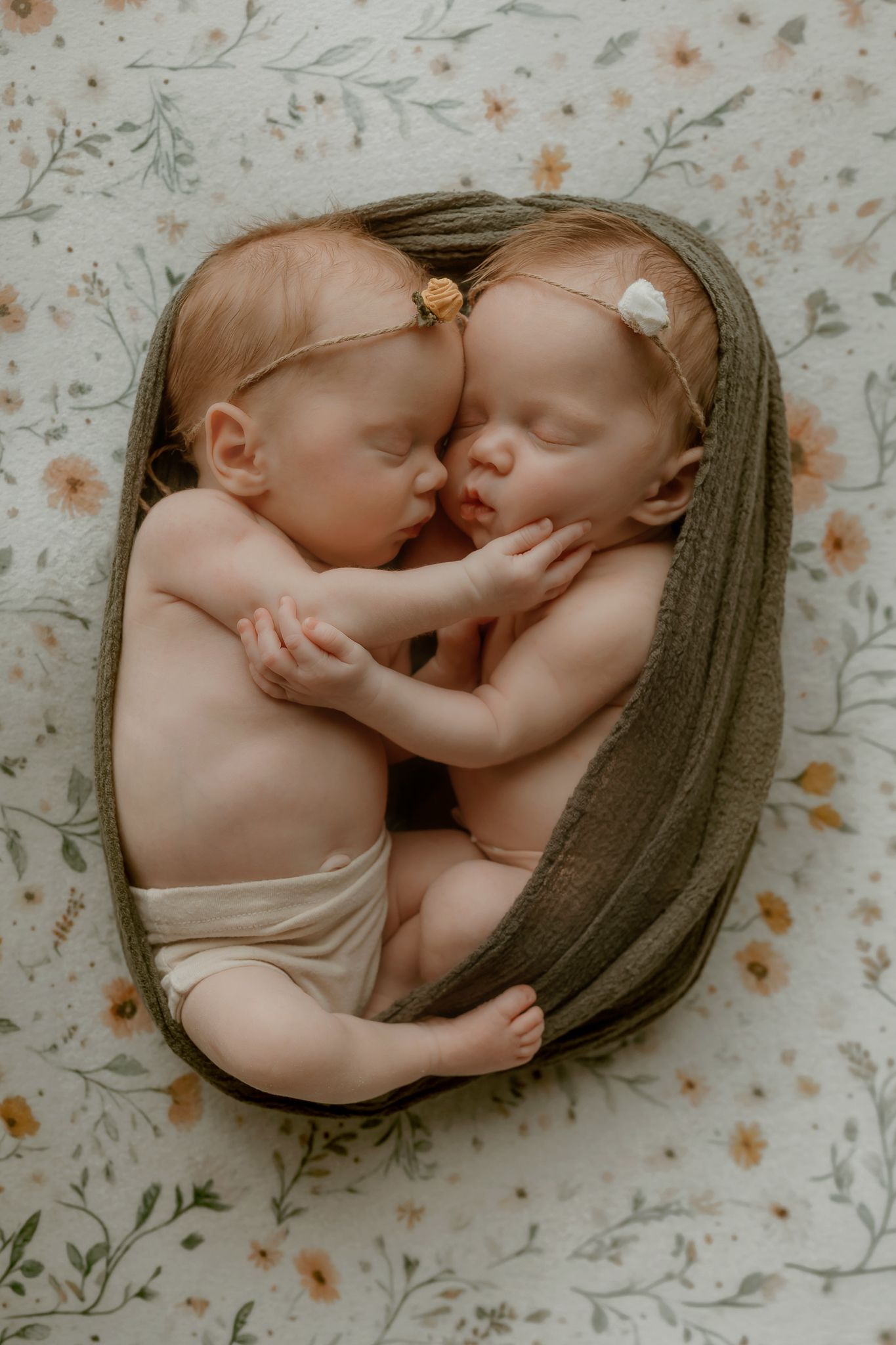 Two sleeping newborn twin babies cuddled together in a bowl, wearing floral headbands, wrapped in olive green fabric.