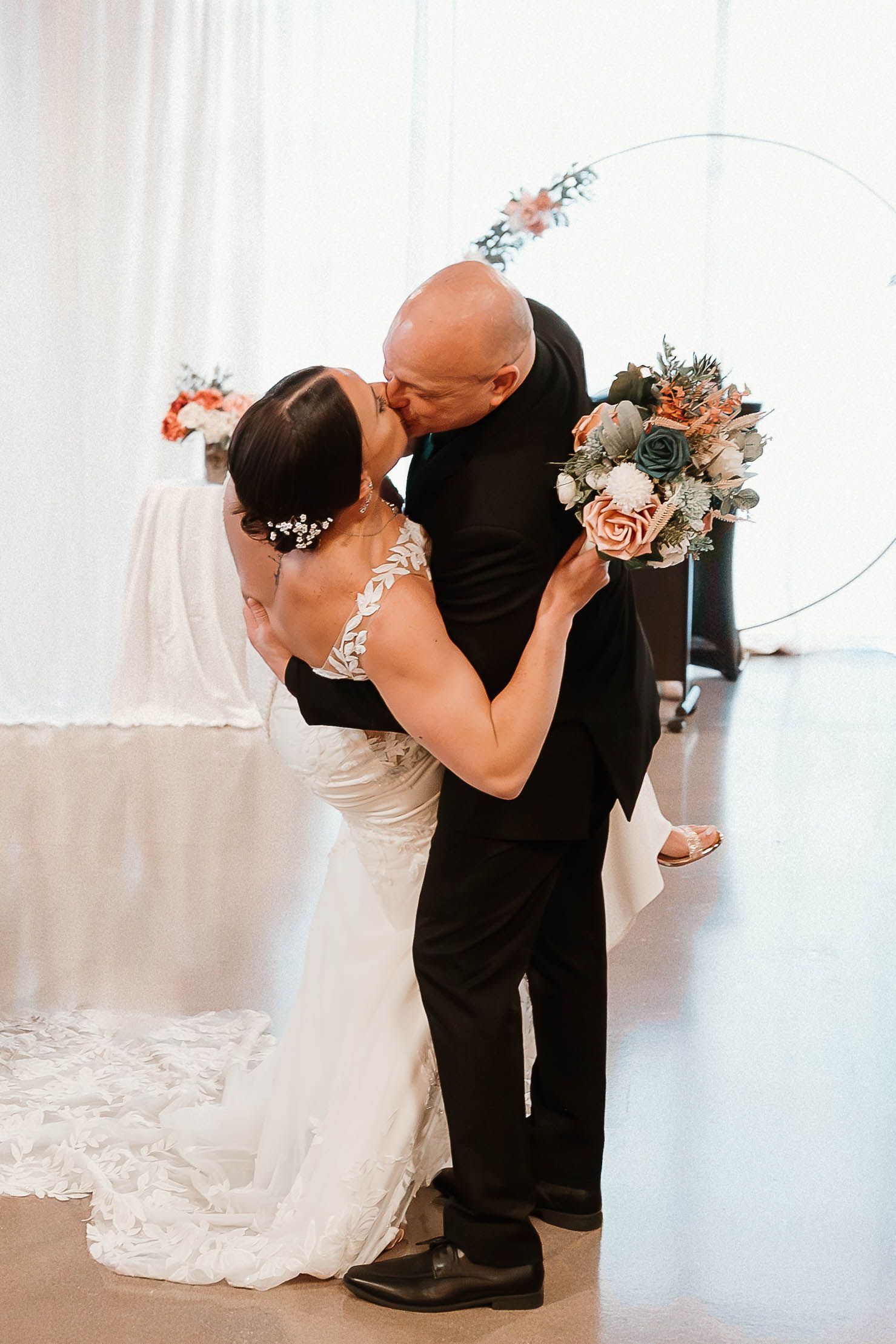 Bride and groom share a romantic first kiss at their wedding ceremony, with a circular floral arch backdrop.