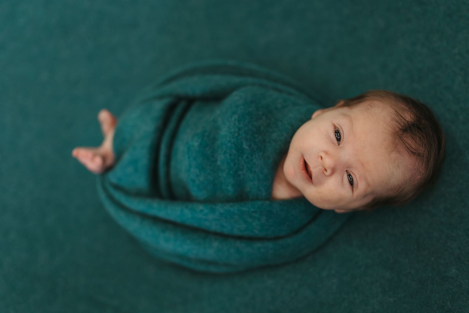 Smiling newborn baby swaddled in teal knit wrap, lying on matching teal backdrop in professional newborn photo.