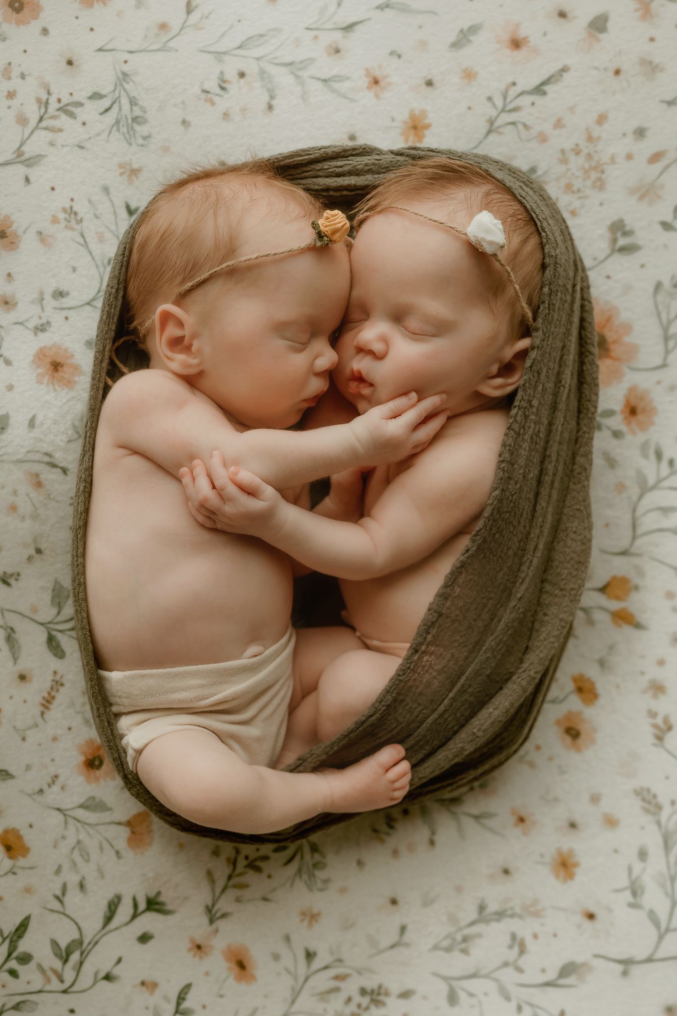 Two sleeping newborn twins cuddle together in a basket wrapped in olive green, wearing floral headbands on a botanical print blanket.