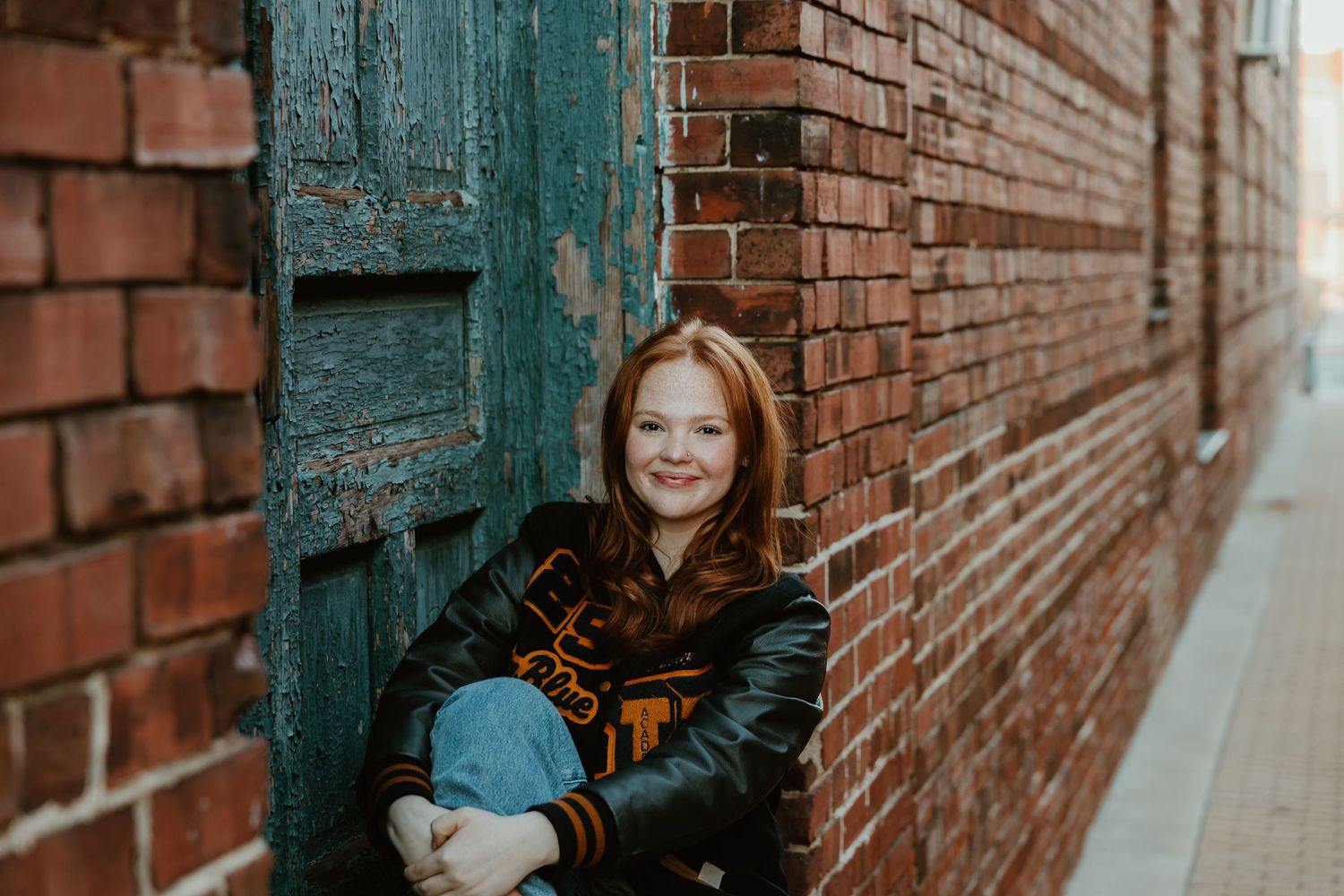 A person with red hair sits against a rustic brick wall next to a weathered teal-colored door.