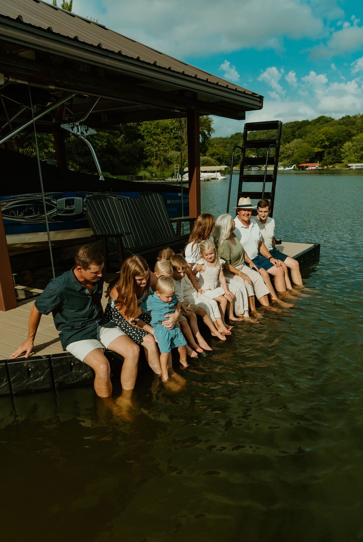 Family group sitting on a lake dock with feet in the water near a boat house on a sunny day.