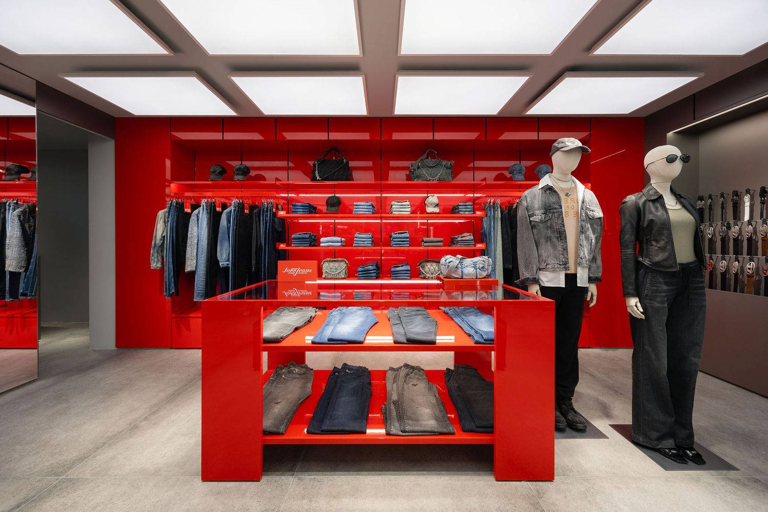 Red retail display shelving with denim jeans, accessories, and two mannequins in a modern clothing store interior.