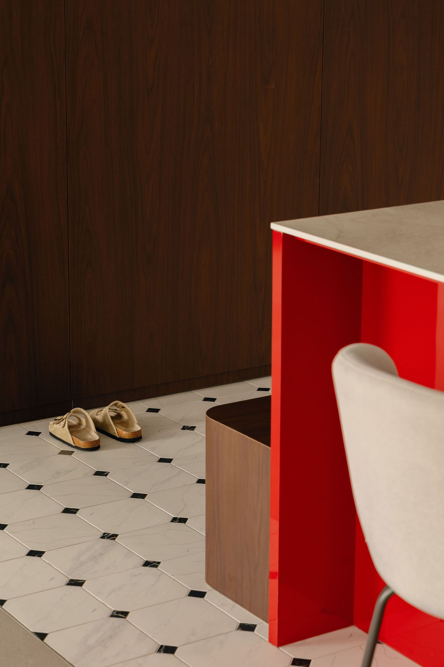 Red modern desk with white chair on black and white tile floor, sandals nearby, wood panel wall backdrop.