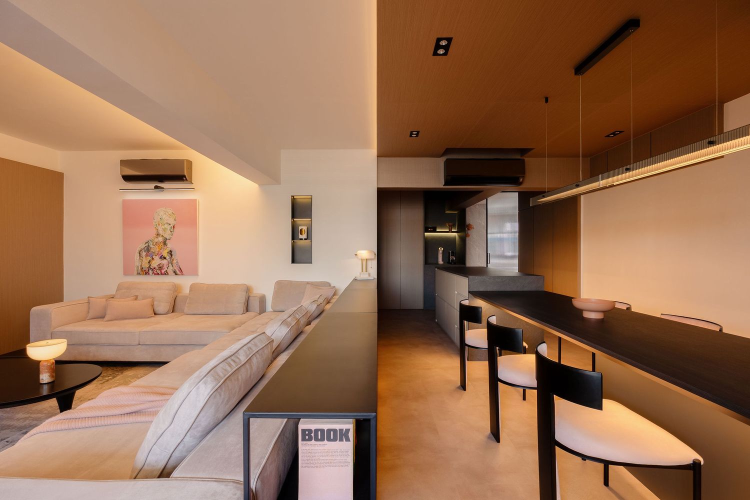 Modern open-plan living room and kitchen with beige sofa, dark bar counter, pendant lights, and warm neutral tones.