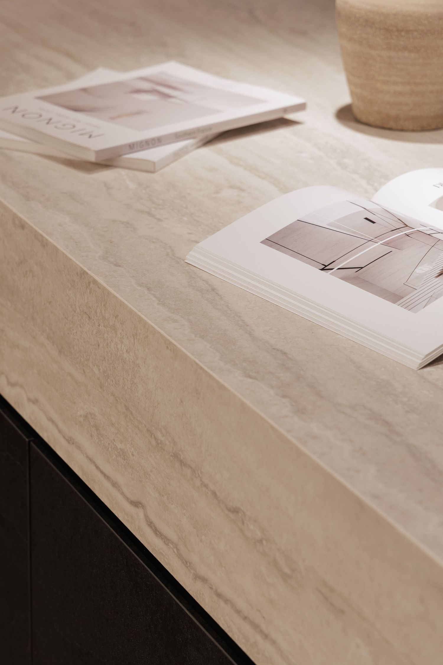 A minimalist scene with open books and a ceramic vessel on a textured marble or stone surface in warm beige tones.