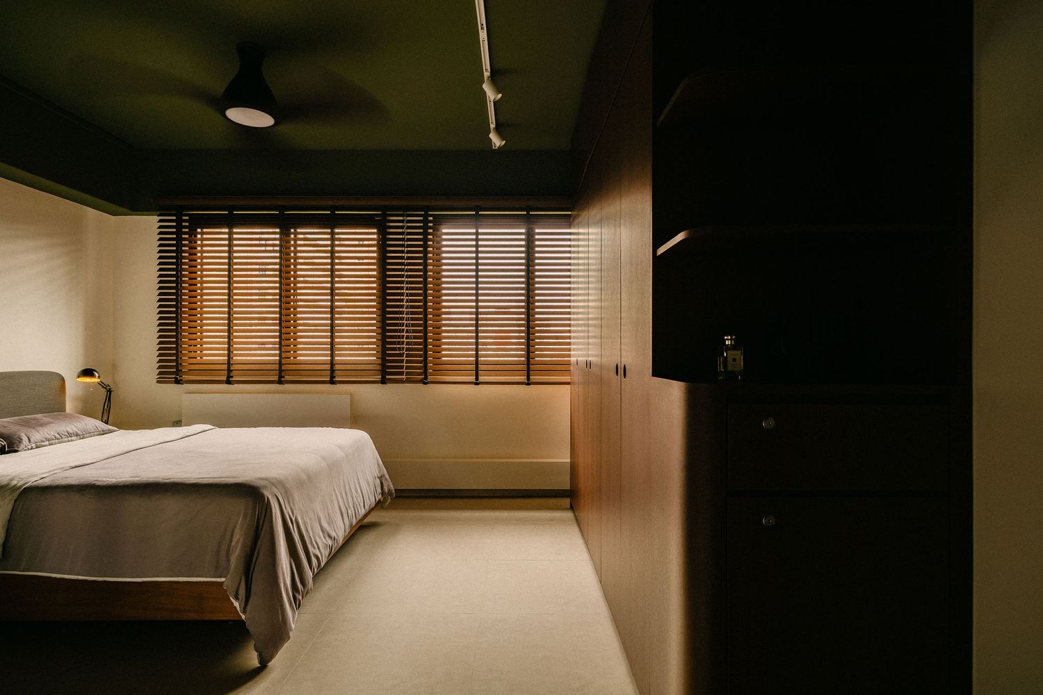 Modern minimalist bedroom with dark green walls, wooden window blinds, a ceiling fan, a neatly made bed and built-in wood cabinetry.