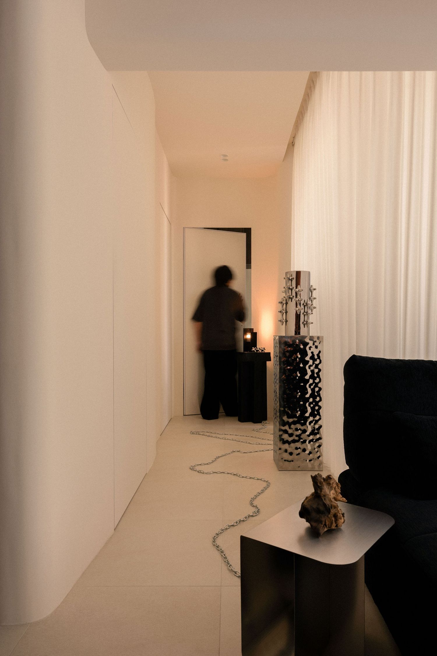 Blurred person walks down a minimalist cream hallway with sheer curtains, a hammered metal vase, a black side table and a decorative rock.