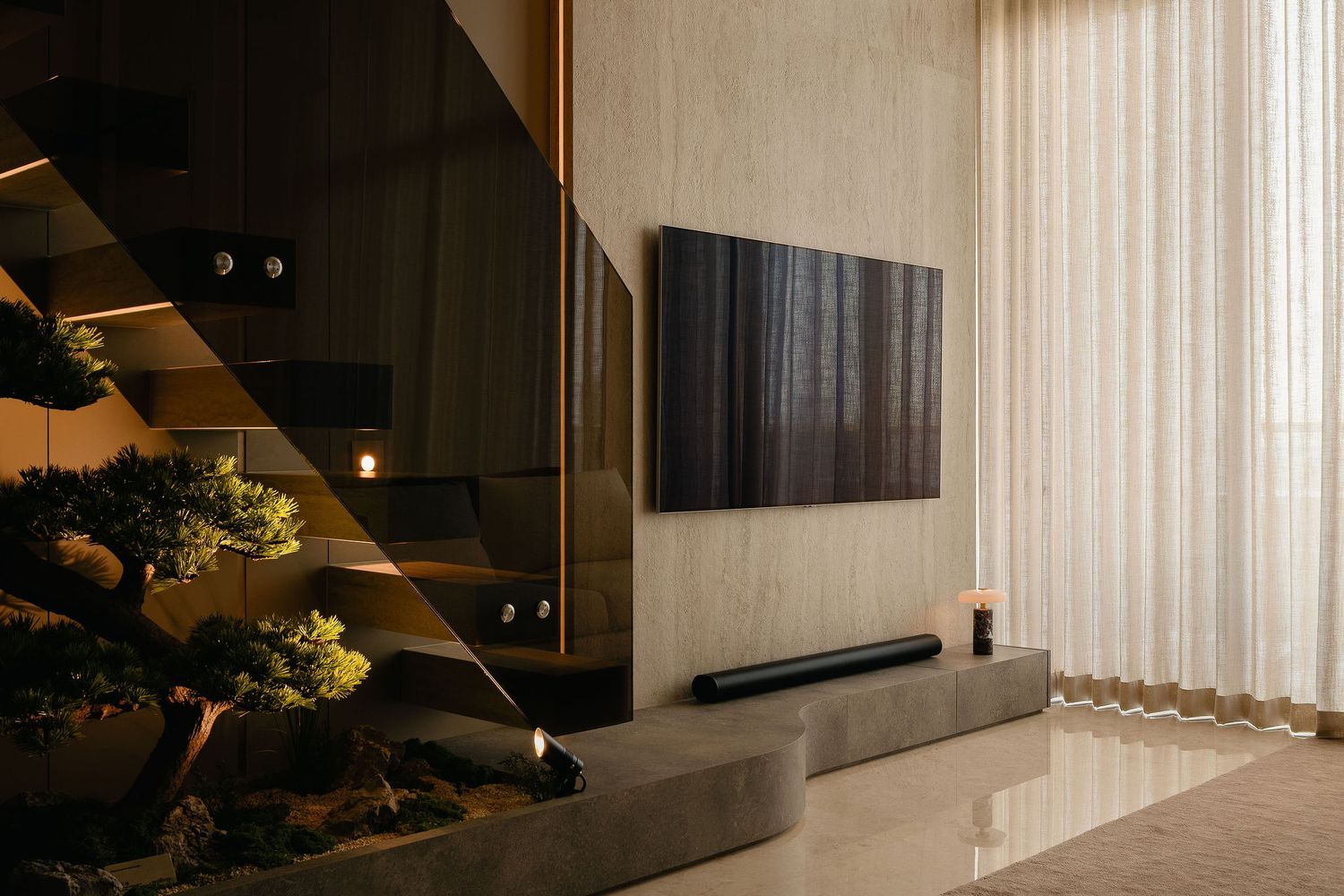 Modern living room with wall-mounted TV, sleek soundbar, sculpted bonsai tree, glass staircase, and sheer curtains in warm neutral tones.