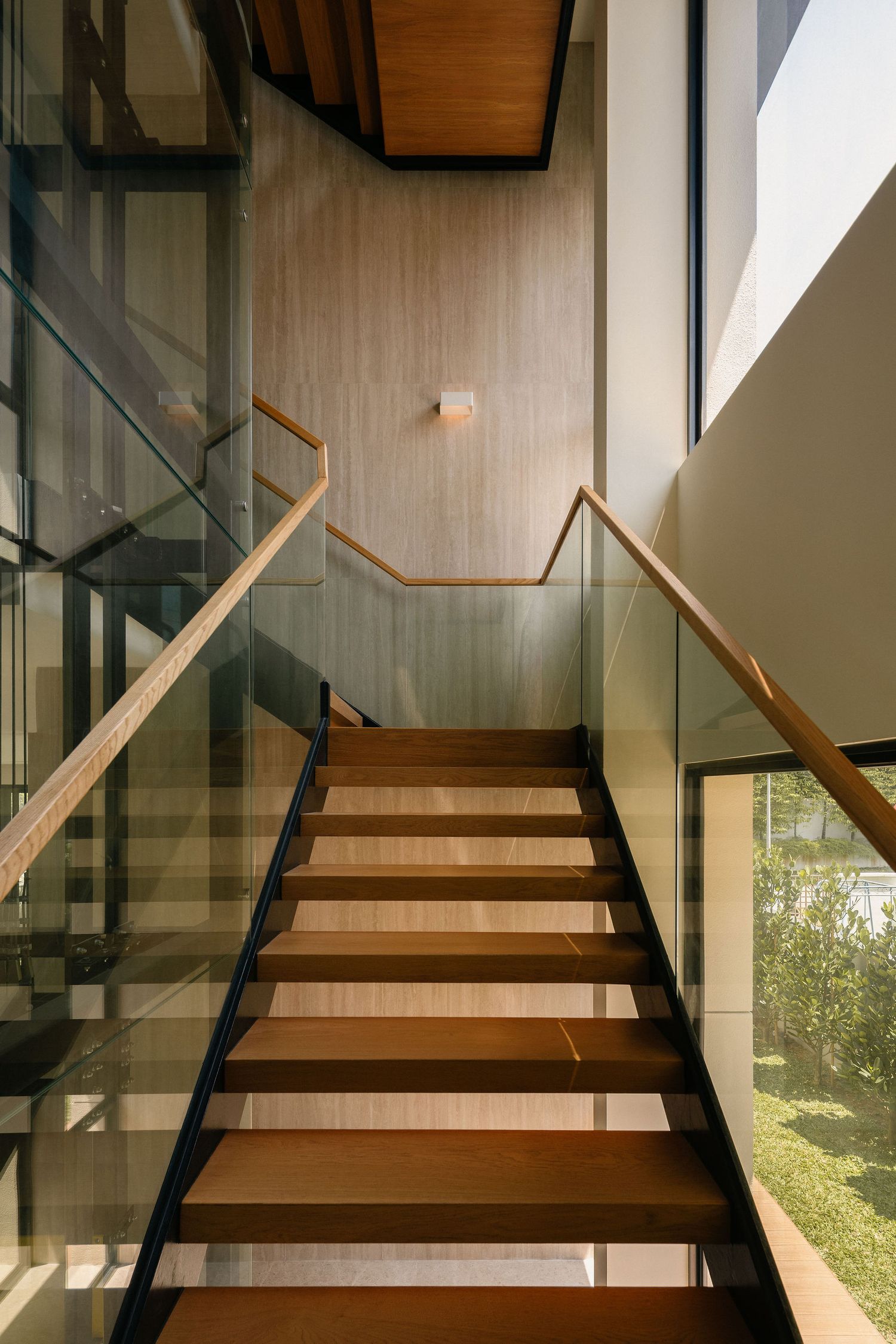 Modern wooden staircase with glass balustrades and wood handrails rising through a bright interior with textured walls and garden views.