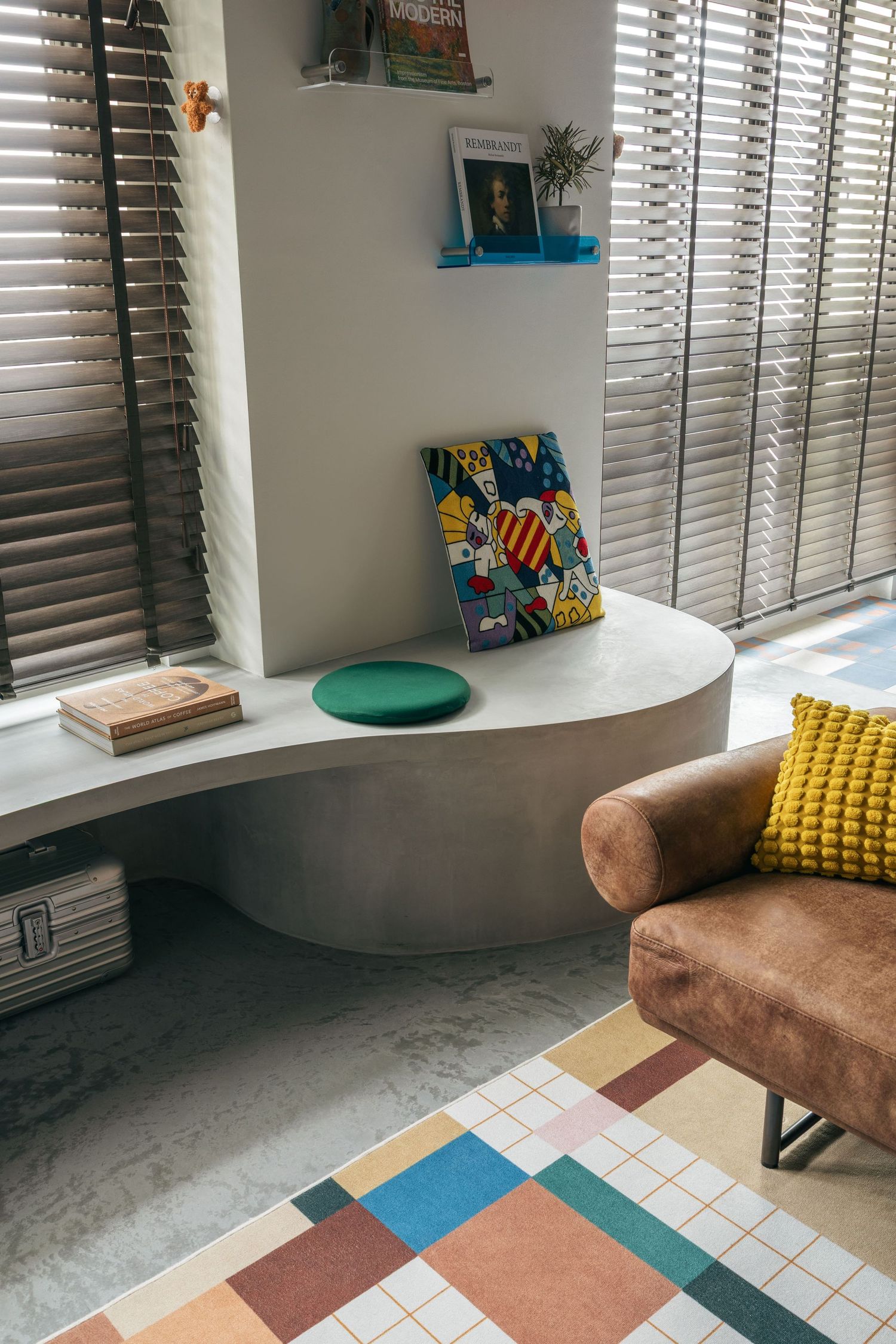 Modern living room corner with curved concrete bench, colorful rug, brown armchair, and decorative shelving by slatted blinds.