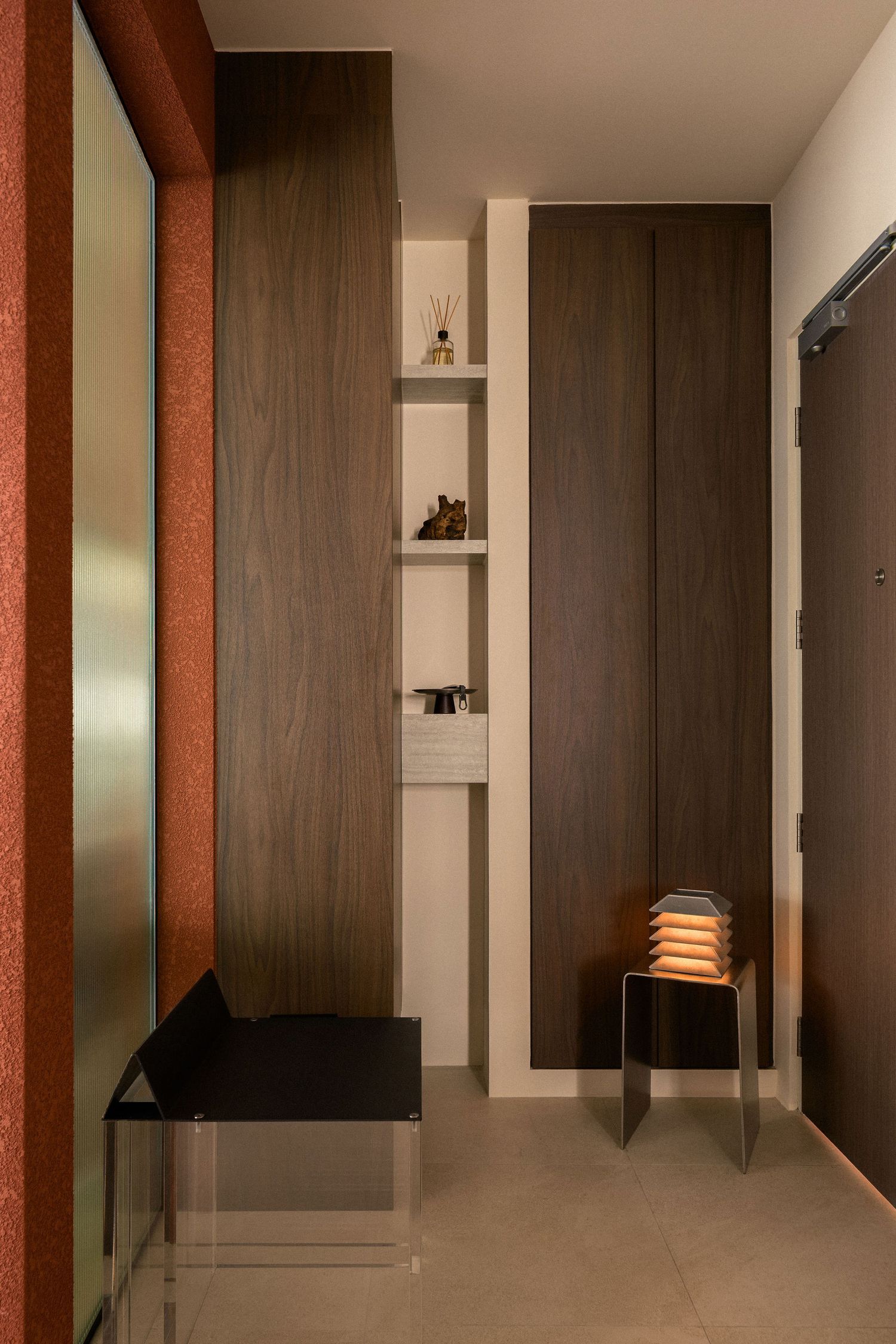Modern entryway with dark wood paneling, built-in shelves with decor, a glass bench, a glowing lantern lamp, and a terracotta accent wall.