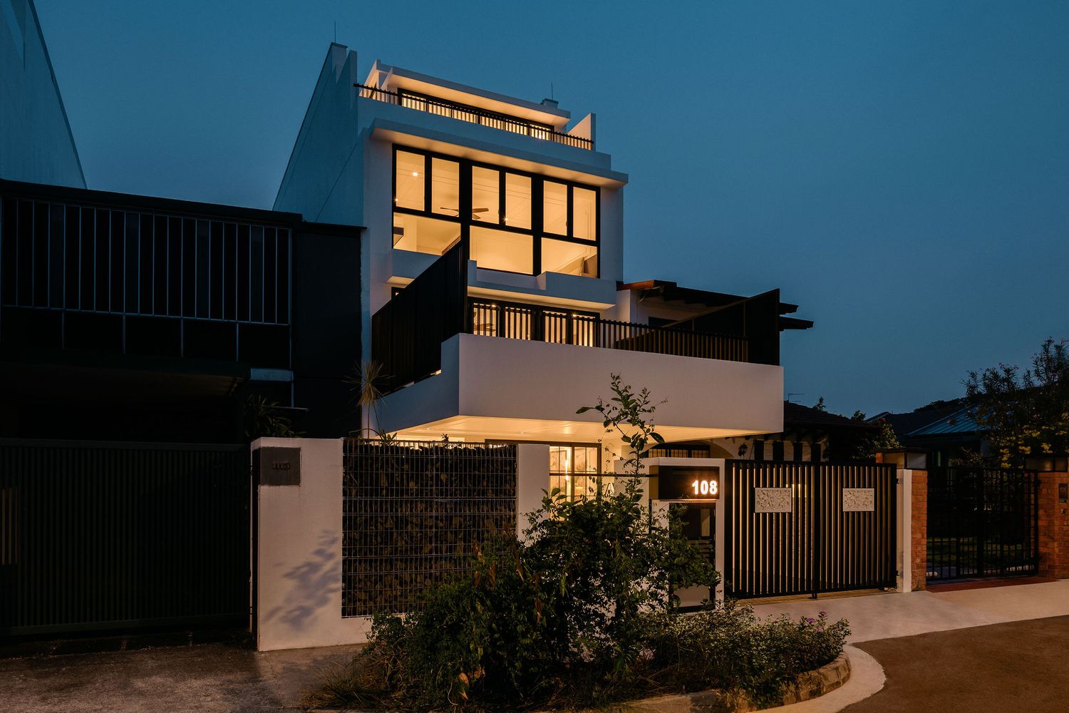 Modern multi-story house with large illuminated windows and a gated entrance, photographed at dusk with landscaped plants in the foreground.
