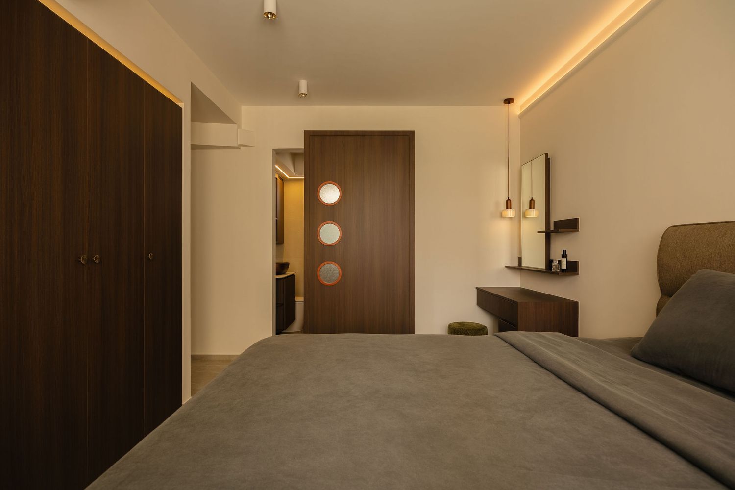 Modern bedroom with a gray bed, dark wood wardrobe, built-in vanity desk with mirror, pendant light and warm cove ceiling lighting.