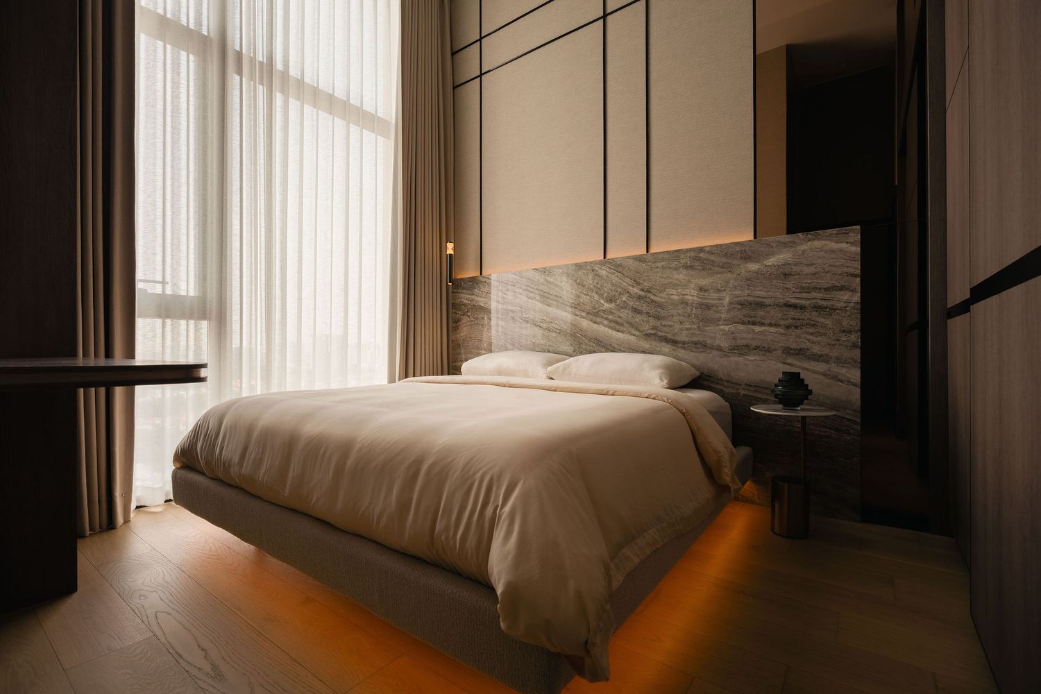 Modern bedroom with a floating bed, warm under-bed lighting, marble headboard wall, beige linens, and floor-to-ceiling windows with sheer cu