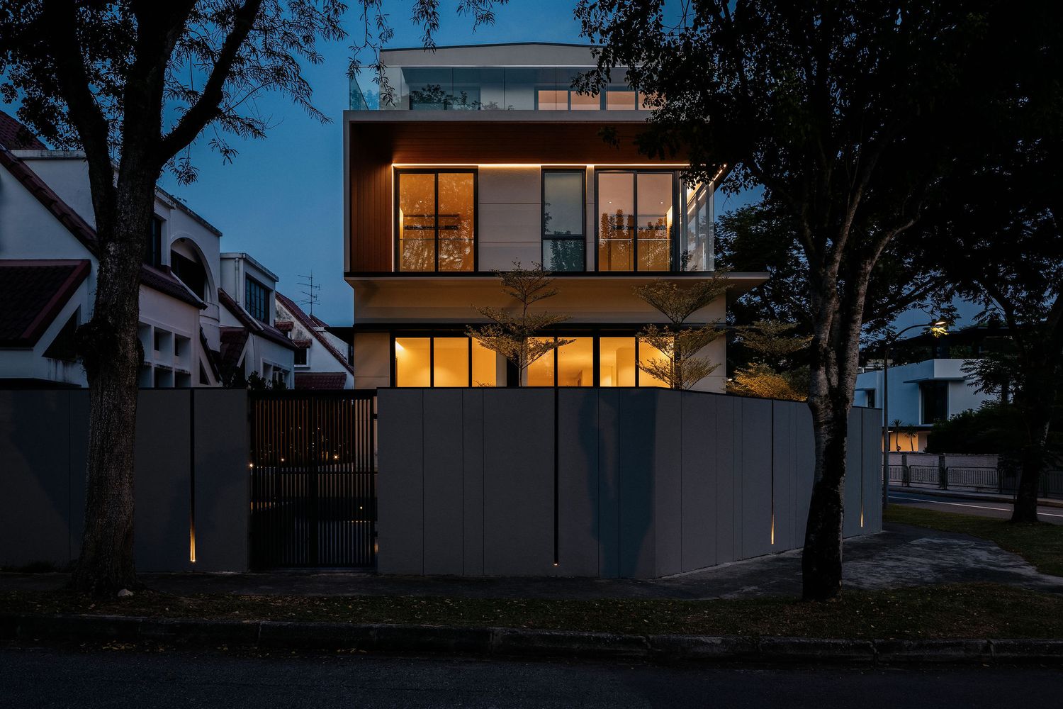 Modern three-story house at dusk with warm glowing windows, wood and concrete facade, tall privacy fence, and mature trees along the street.