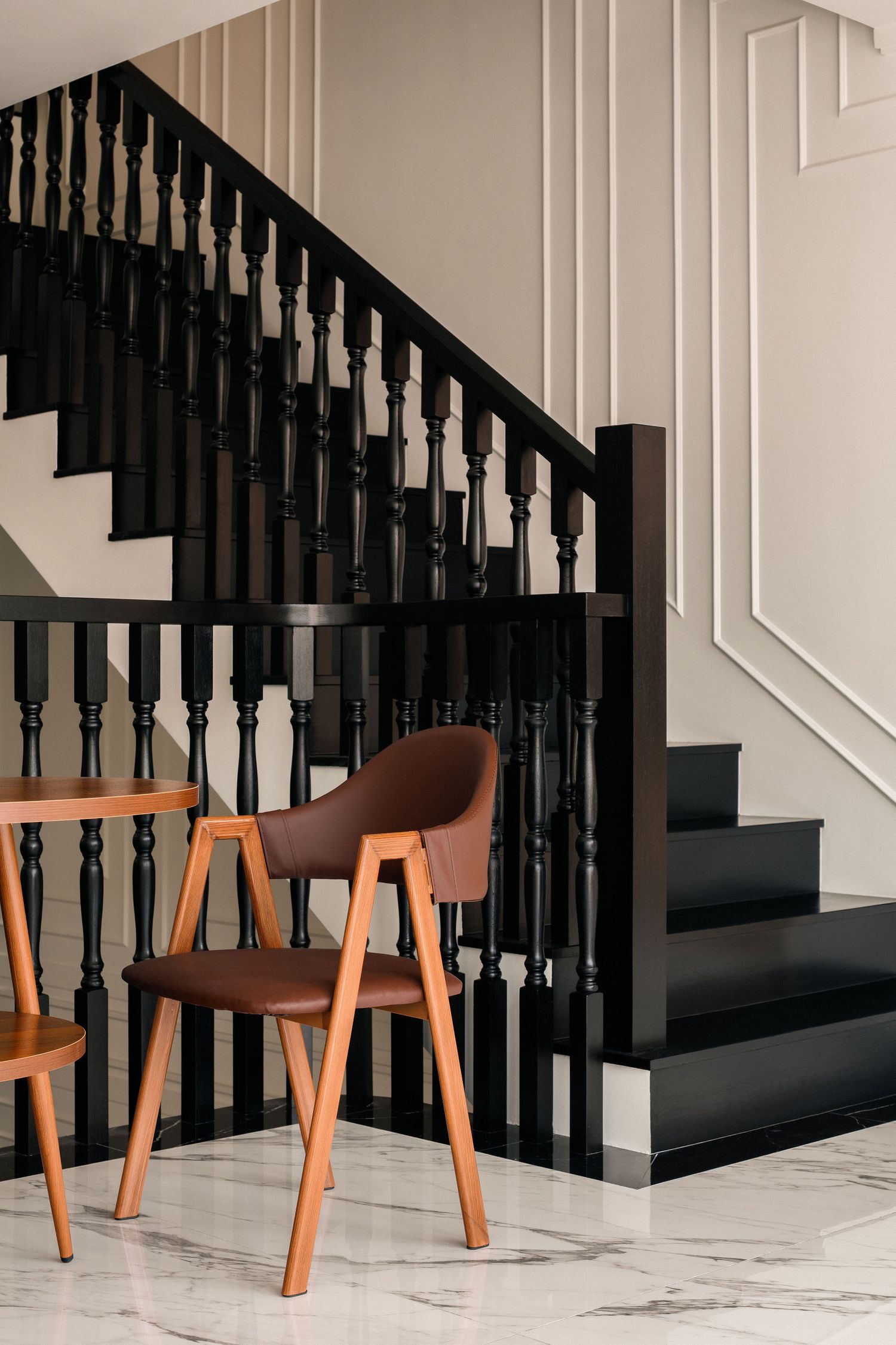 Brown leather dining chair with wooden legs beside a dark wood staircase with black balusters and white wall paneling on a marble floor.