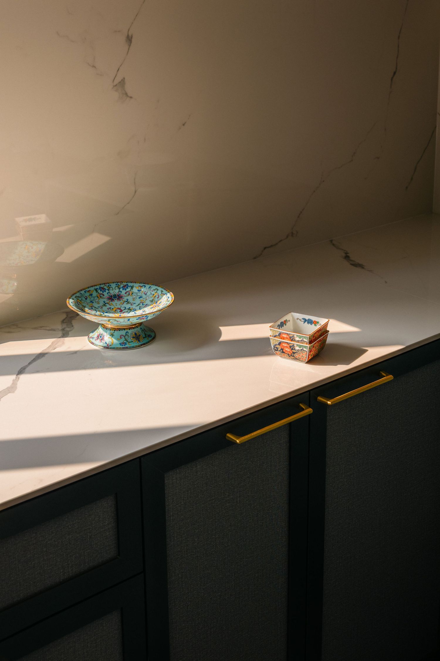 Sunlit white marble countertop with a vintage painted pedestal dish and small porcelain boxes above dark cabinets with brass handles.