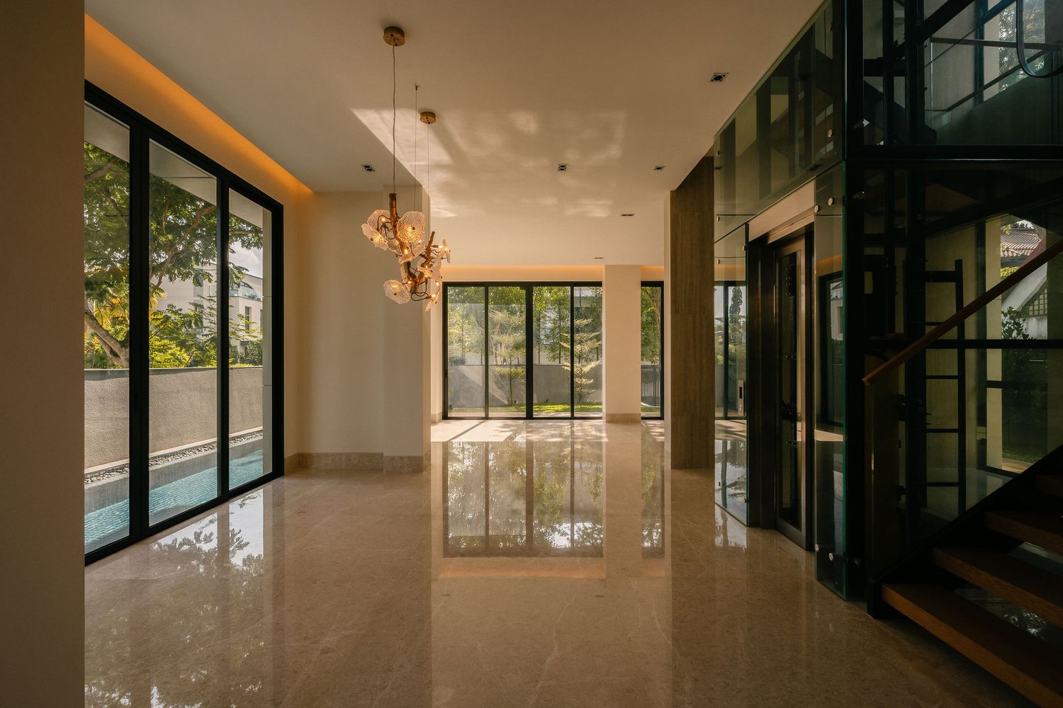 Spacious modern interior with polished marble floors, floor-to-ceiling glass windows, a pendant chandelier, and a black steel staircase.